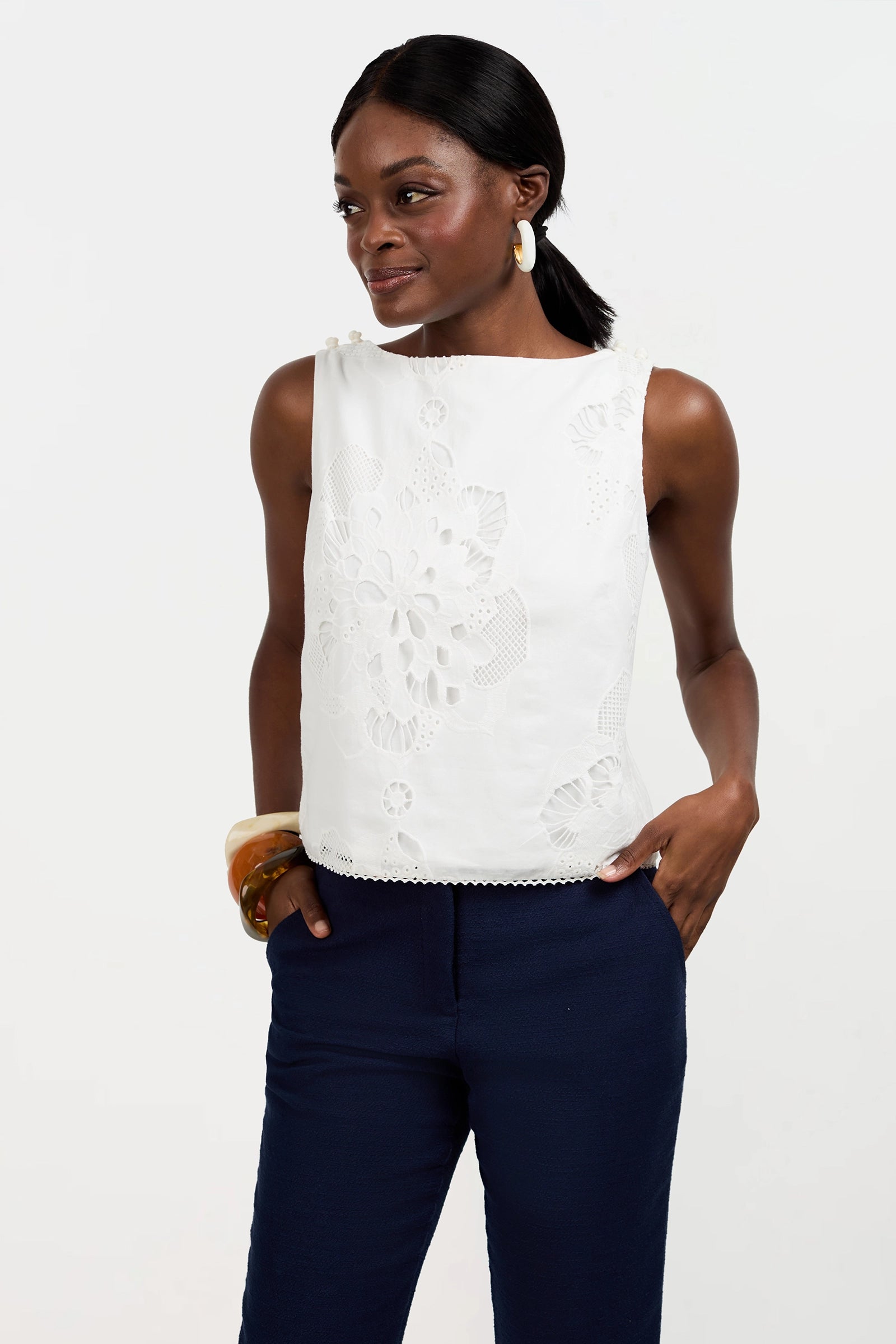 A woman with dark hair in a low ponytail wears the sleeveless white embroidered Alessandra Top and dark blue pants, accessorized with large hoop earrings and a bangle, standing with her hands in her pockets against a plain white background.