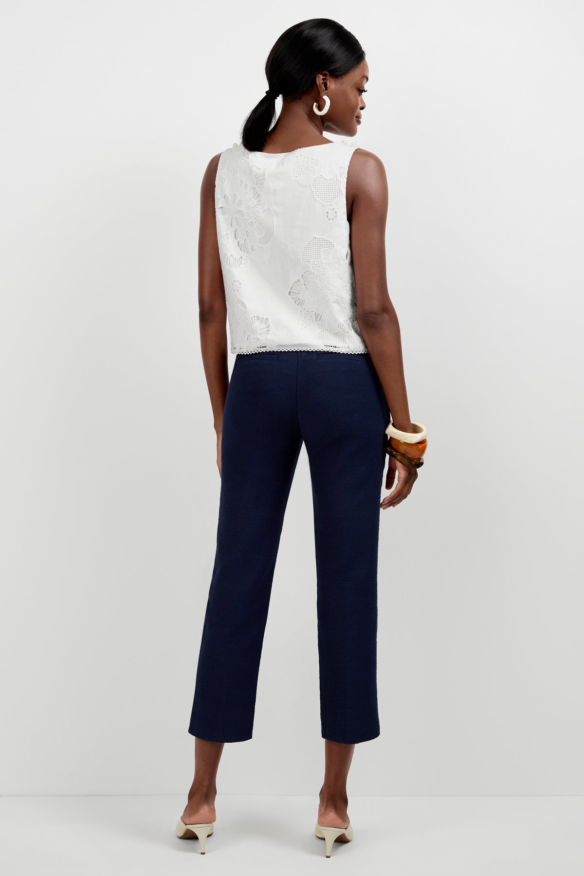 A woman faces away against a plain white background, wearing the Diffuse Pant in navy blue with a tulip hem, a sleeveless white lace top, cream low-heeled shoes, large hoop earrings, and chunky bracelets.