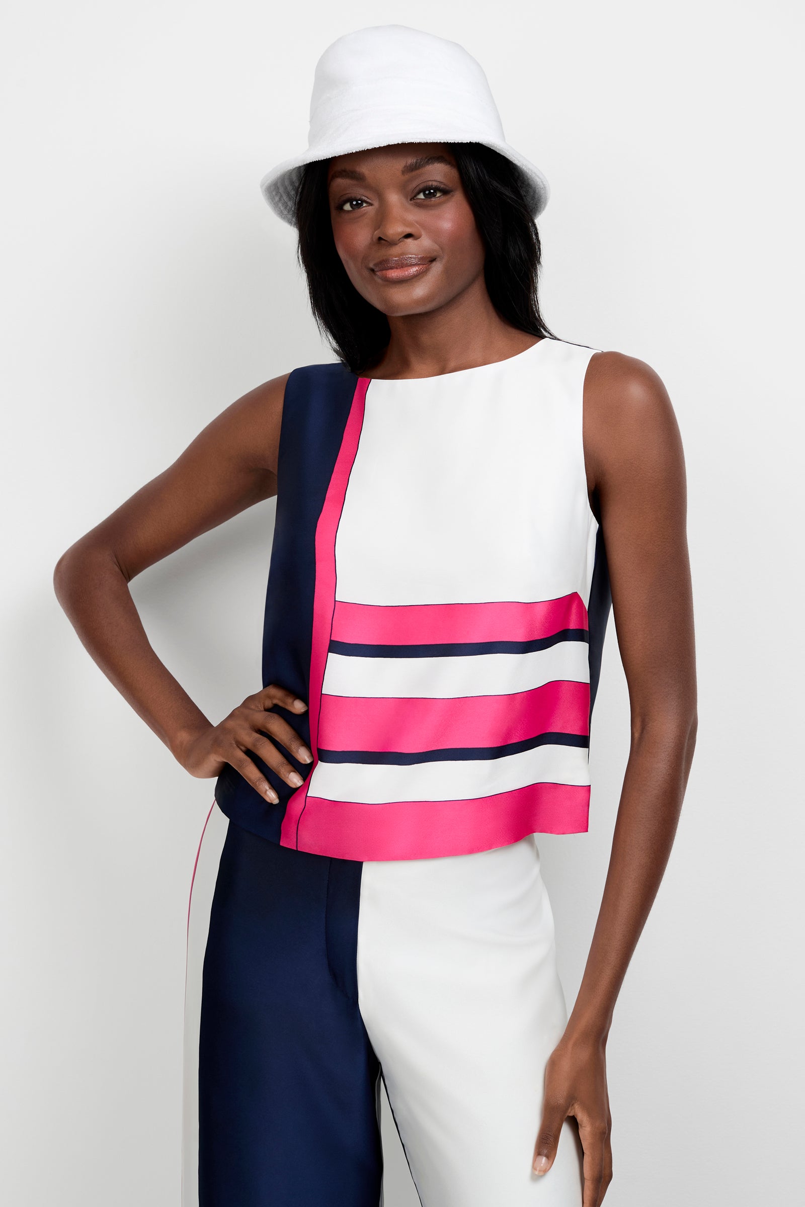A woman smiles with one hand on her hip, wearing the Cadorna Linee Sleeveless Top—pink and navy color-blocked with stripes—paired with split-color pants and a white bucket hat, posed against a plain white background.