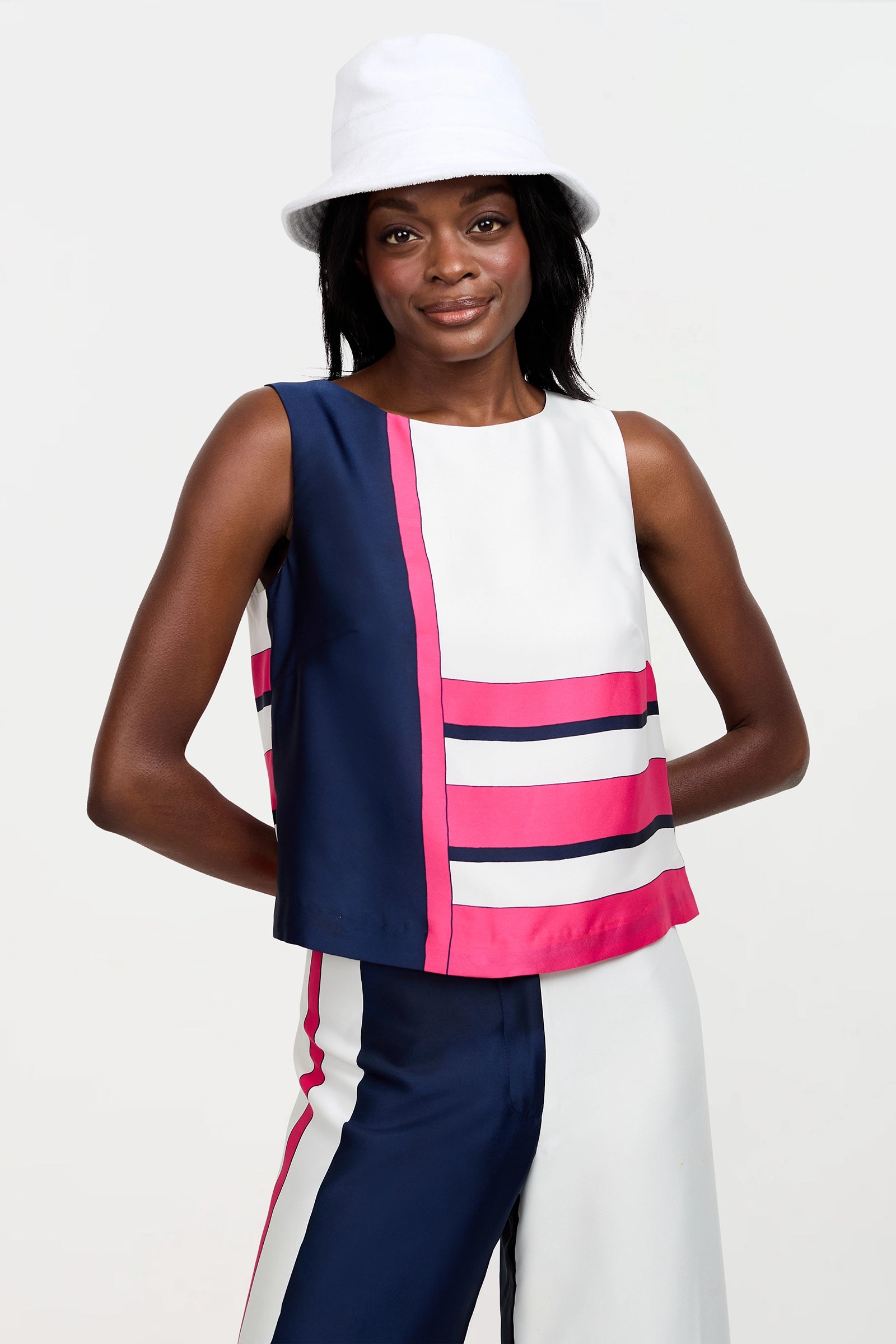 A woman wearing the Cadorna Linee Sleeveless Top and matching pants with bold navy blue, white, and pink color blocks, plus a white bucket hat, stands against a light background and smiles softly.