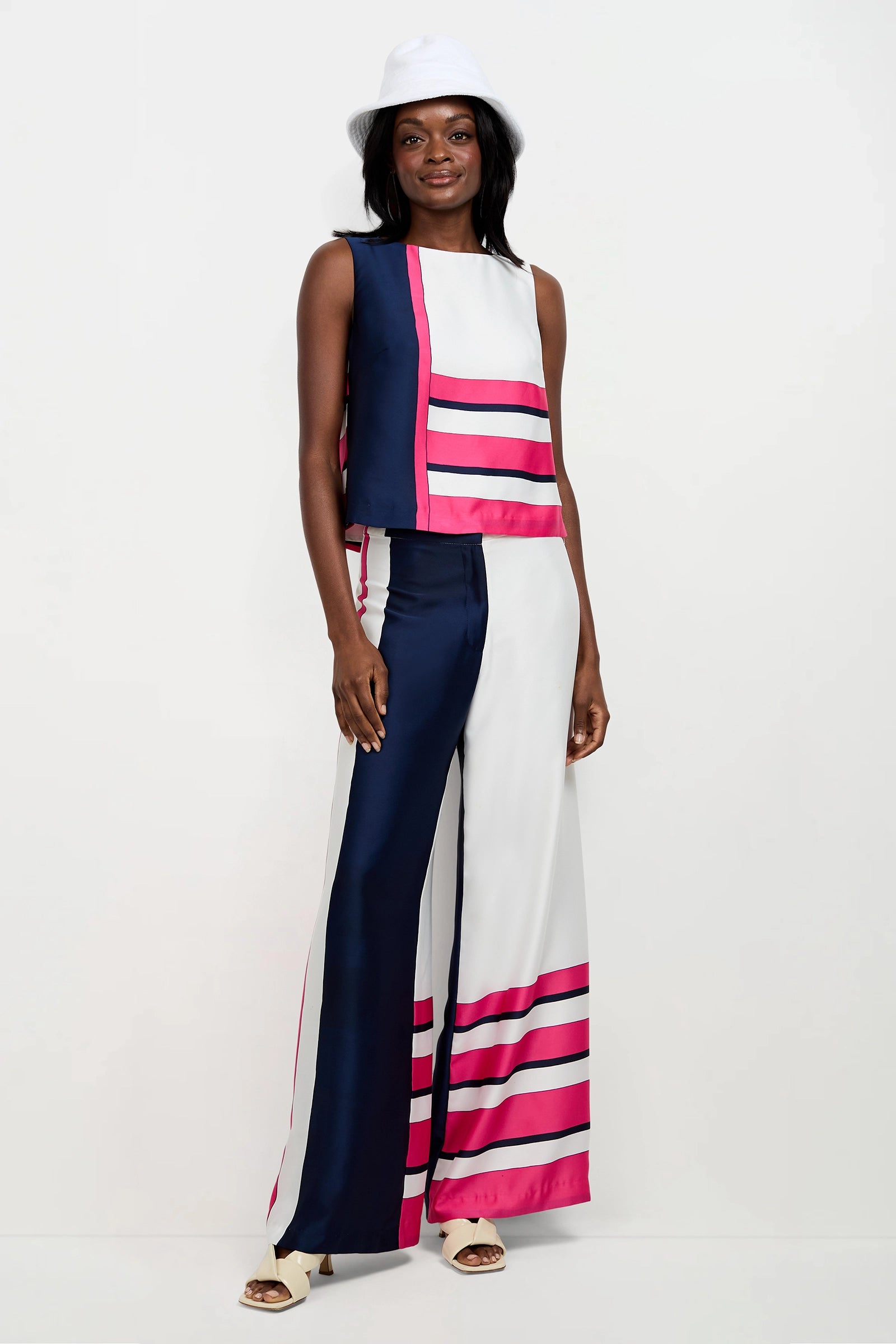 A woman wears the Cadorna Linee Sleeveless Top, featuring navy blue, white, and pink color blocks and stripes, styled with wide-leg pants, a white hat, and open-toe sandals against a plain white background.