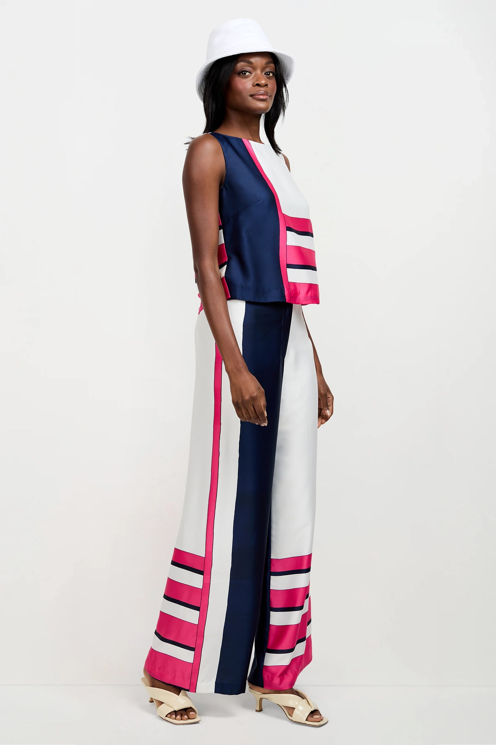 A woman wears the Cadorna Linee Sleeveless Top in navy, white, and pink with matching wide-leg pants, styled with a white hat and heeled sandals. She stands sideways against a plain white background.