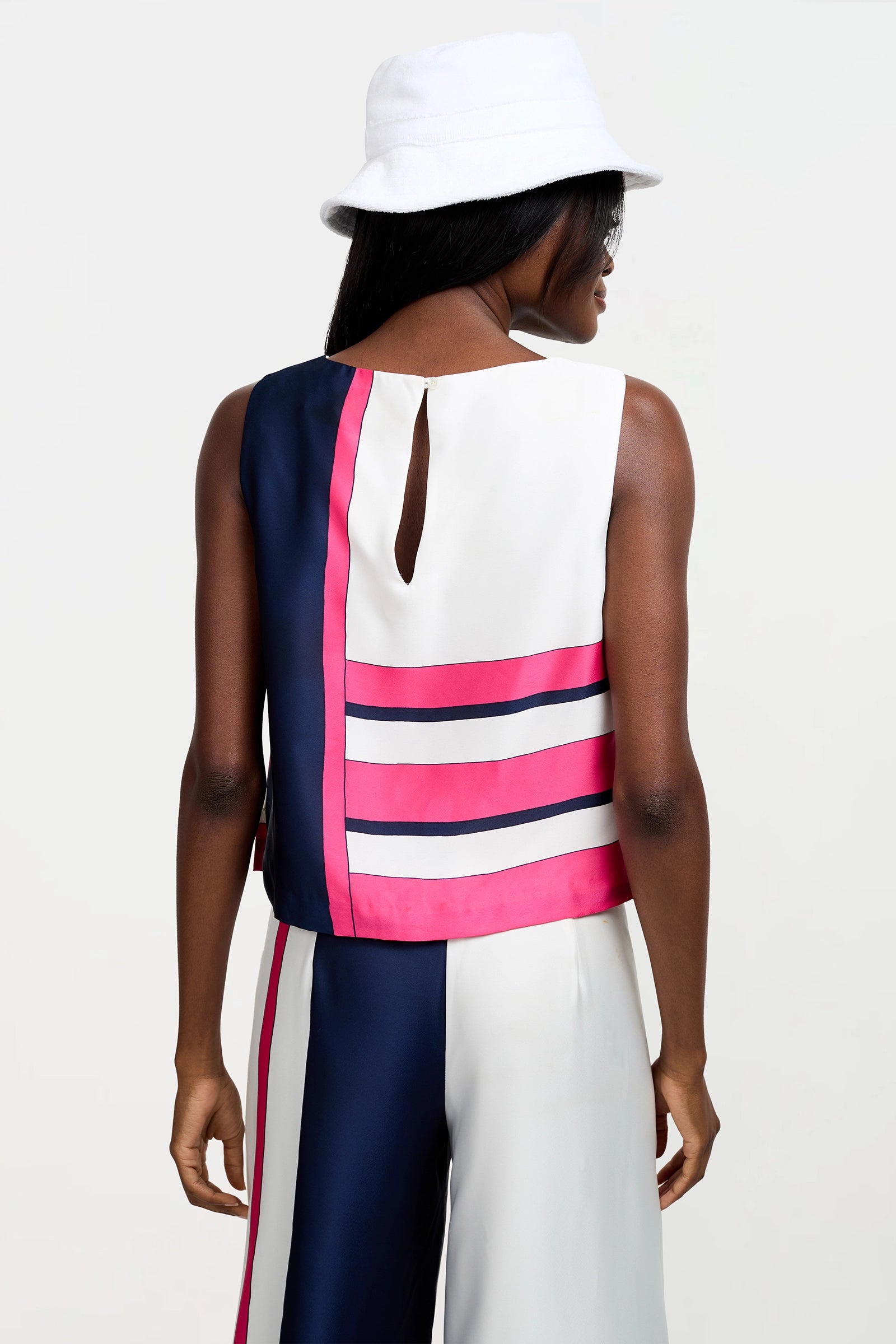 A woman faces away wearing the Cadorna Linee Sleeveless Top with navy, white, and pink stripes, matching pants, and a white bucket hat against a plain background.
