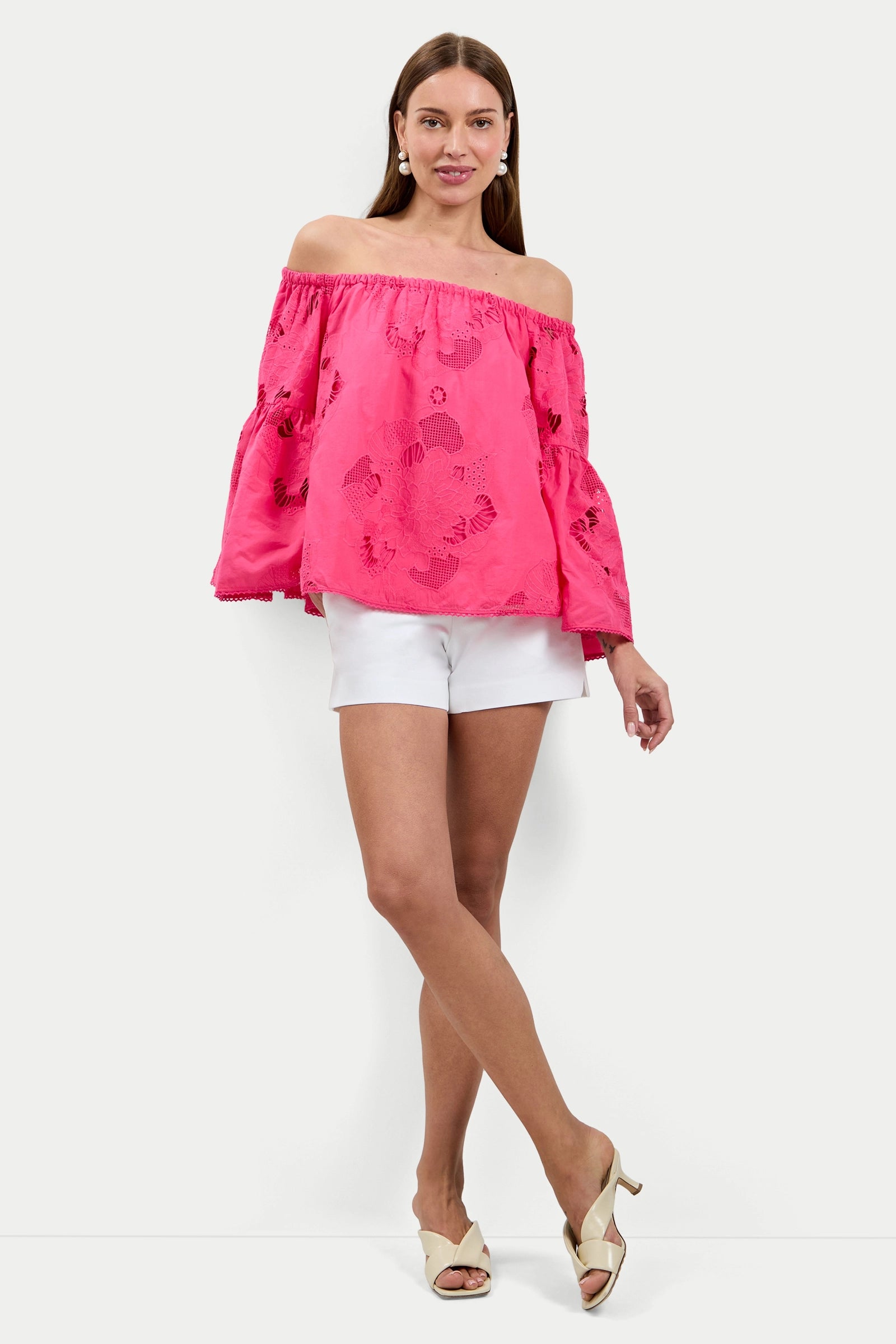 A woman smiles while posing against a white background, wearing the Perry Off-Shoulder Top in bright pink with bell sleeves, paired with white shorts and cream heeled sandals.