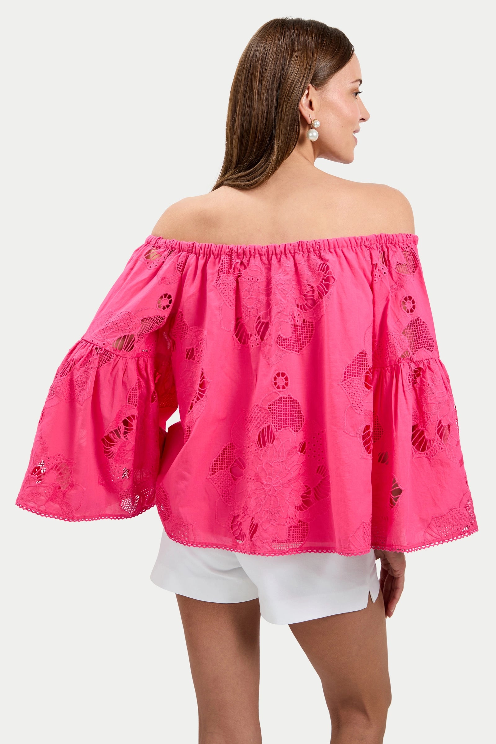 A woman with long brown hair wears the Perry Off-Shoulder Top in bright pink with embroidery and wide flared sleeves, paired with white shorts and pearl earrings. She is facing away against a plain white background.