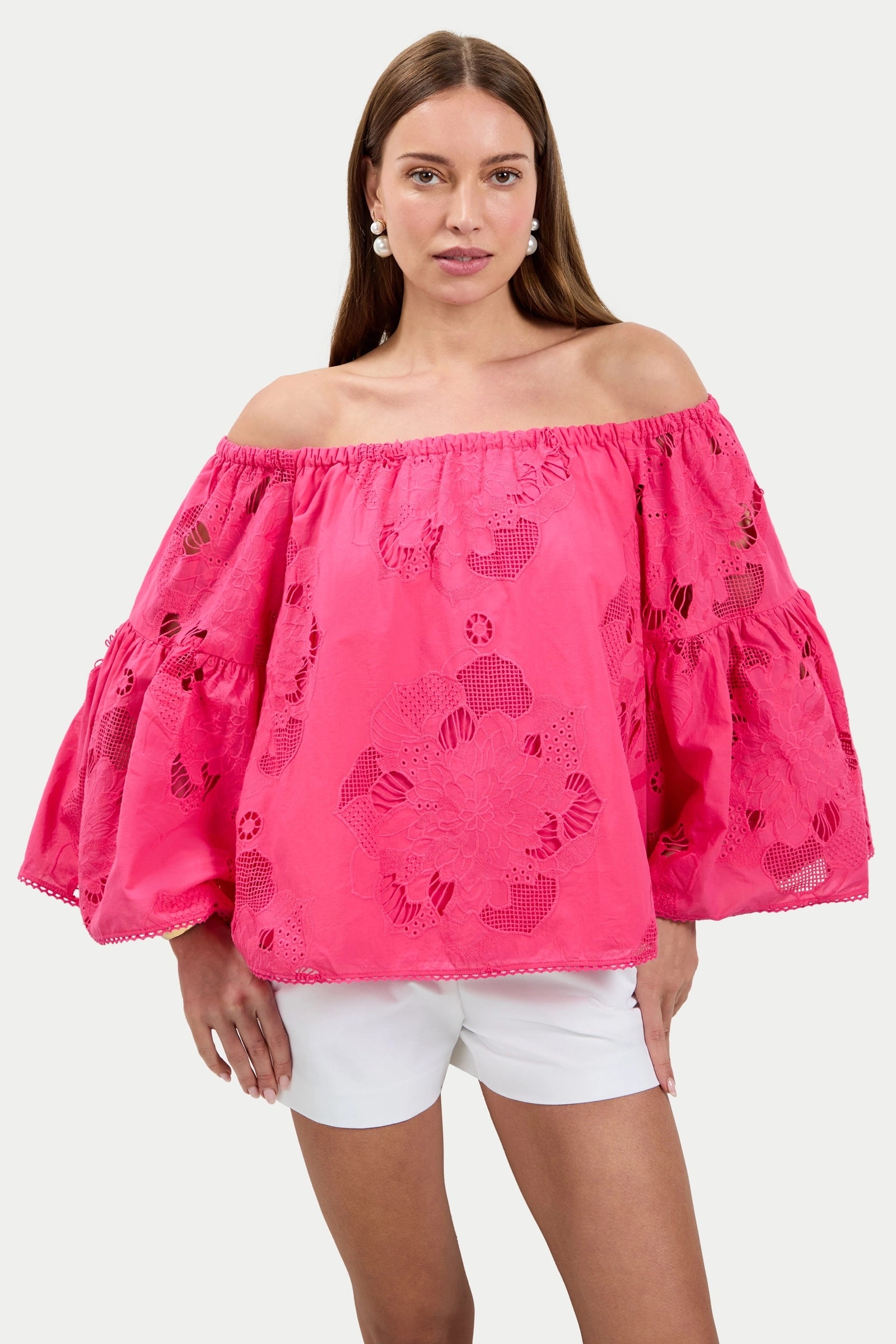 A woman models the Perry Off-Shoulder Top in bright pink with floral cut-out details and white shorts, posing with one hand on her hip against a plain background.