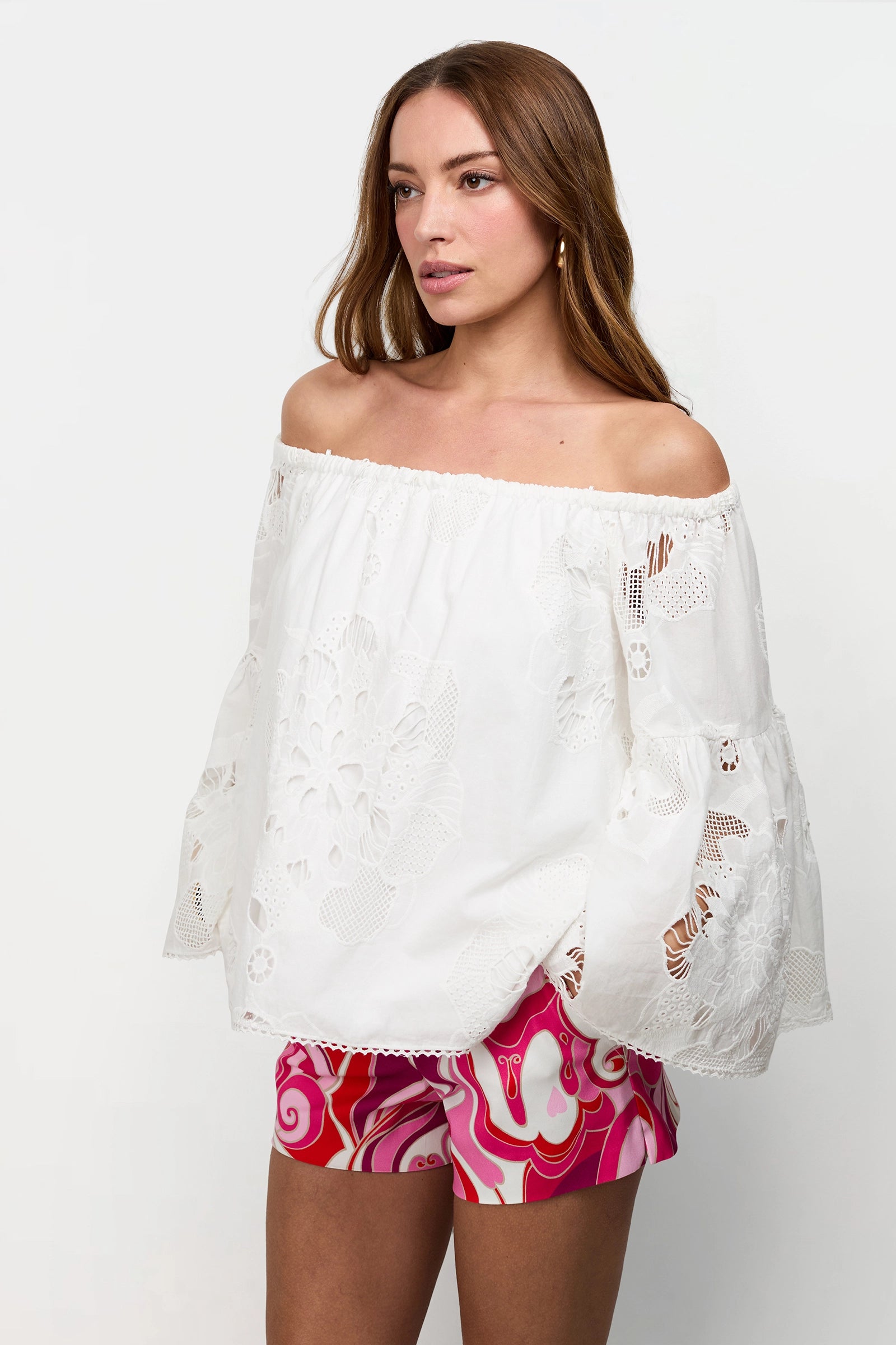 A woman models the Perry Off-Shoulder Top, a white lace-patterned blouse with wide sleeves, paired with pink patterned shorts, standing against a plain white background.