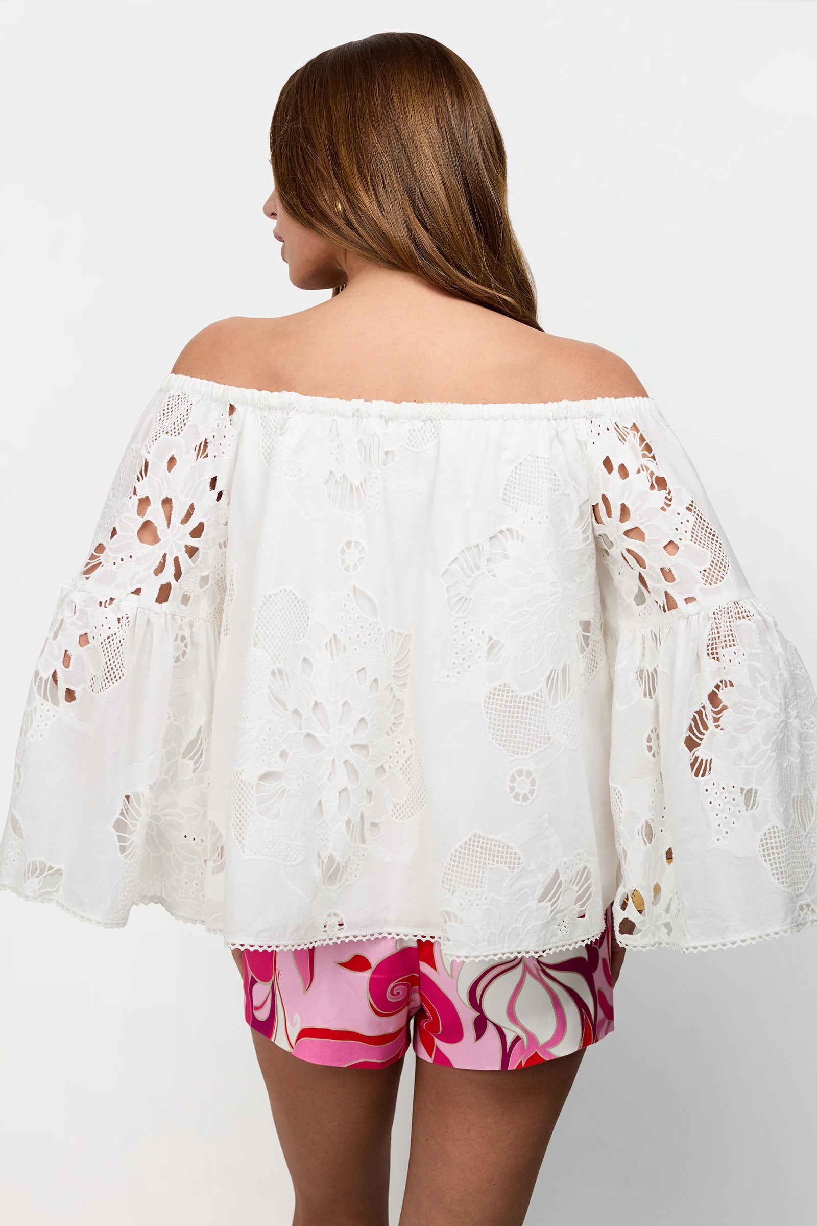 A woman with long brown hair is seen from behind, wearing the Perry Off-Shoulder Top—a white lace blouse with wide sleeves—paired with pink patterned shorts against a plain white background.