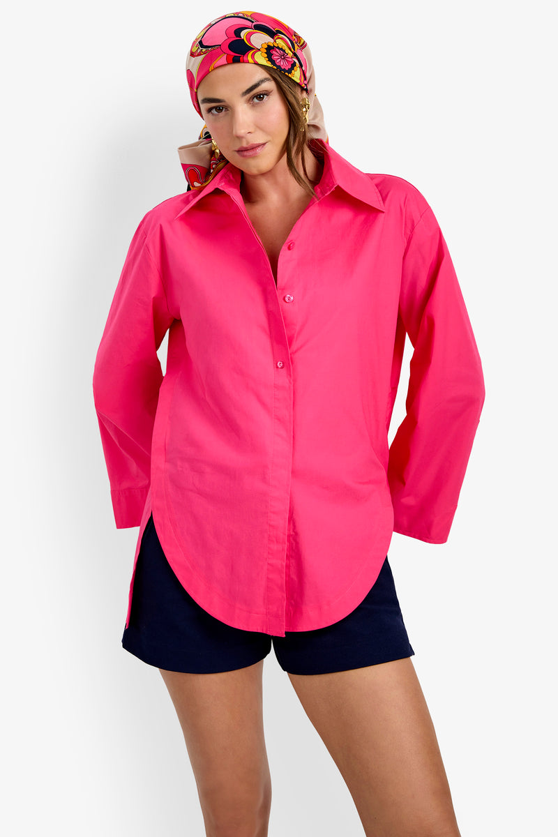 A woman wearing the Olona Swing Top in bright pink, paired with dark shorts and a colorful floral headscarf, stands against a plain white background.