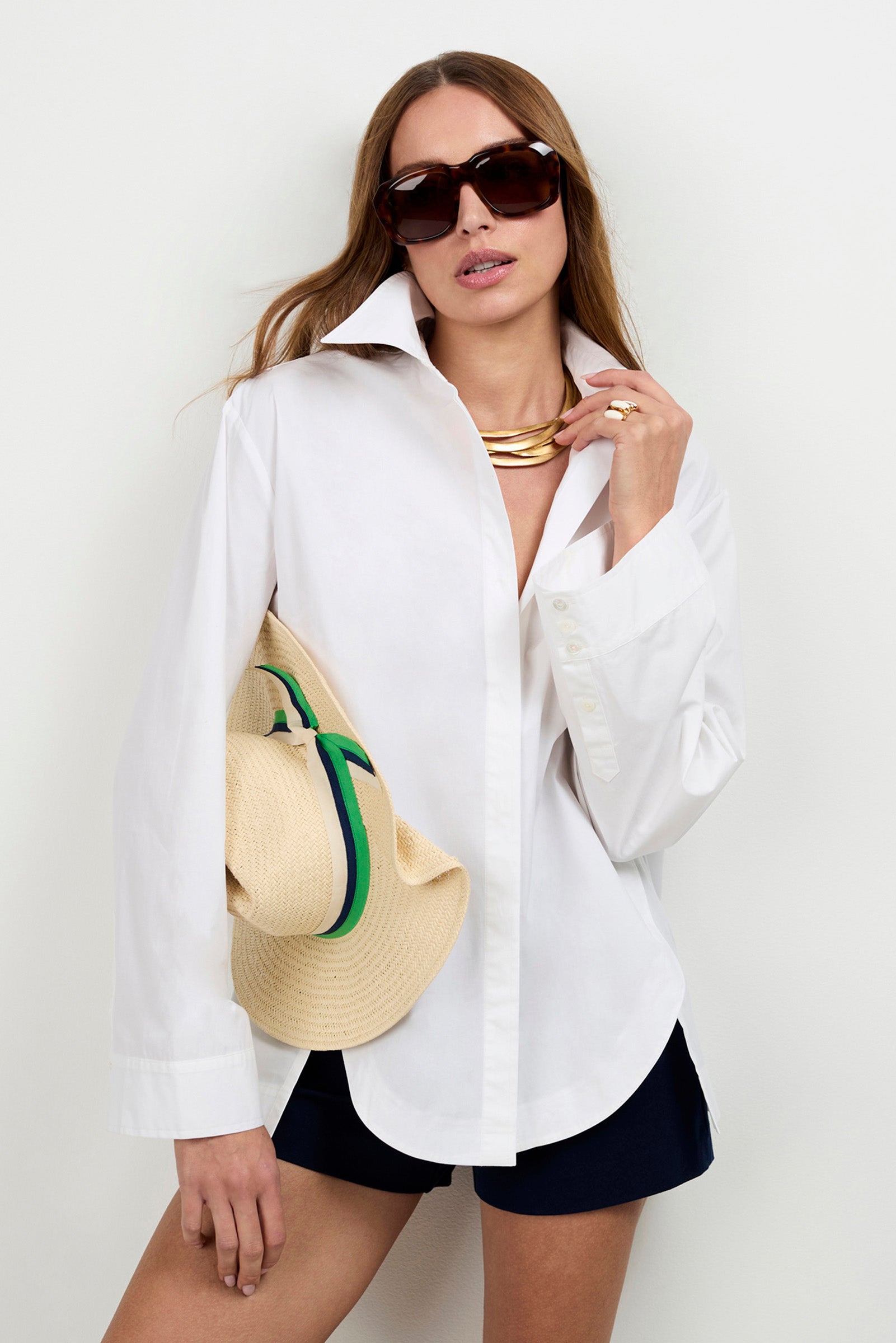 A woman wears the Olona Swing Top, black shorts, large sunglasses, and a gold necklace while holding a straw hat trimmed in green and blue. She poses against a plain white background with long hair and a gold ring.