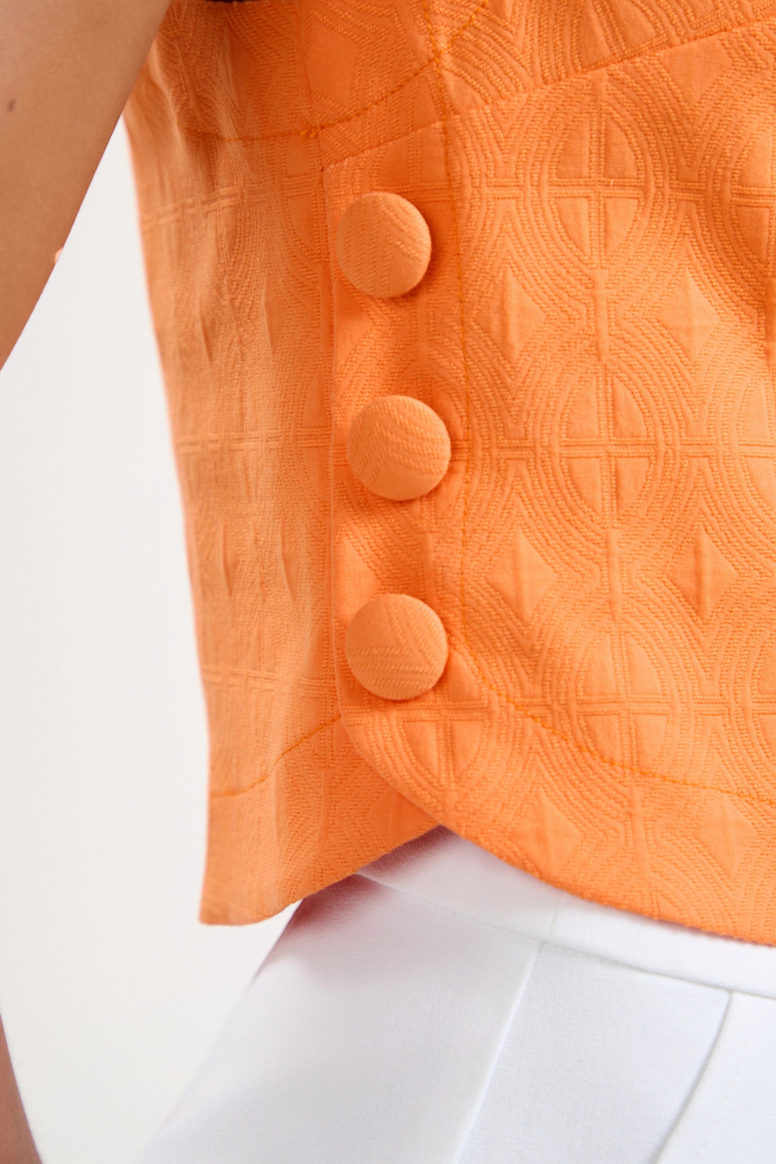 Close-up of a person wearing the Siara Button Side Top—an orange textured sleeveless top with three large side buttons—paired with white high-waisted pants. Only part of the torso and arm are visible.
