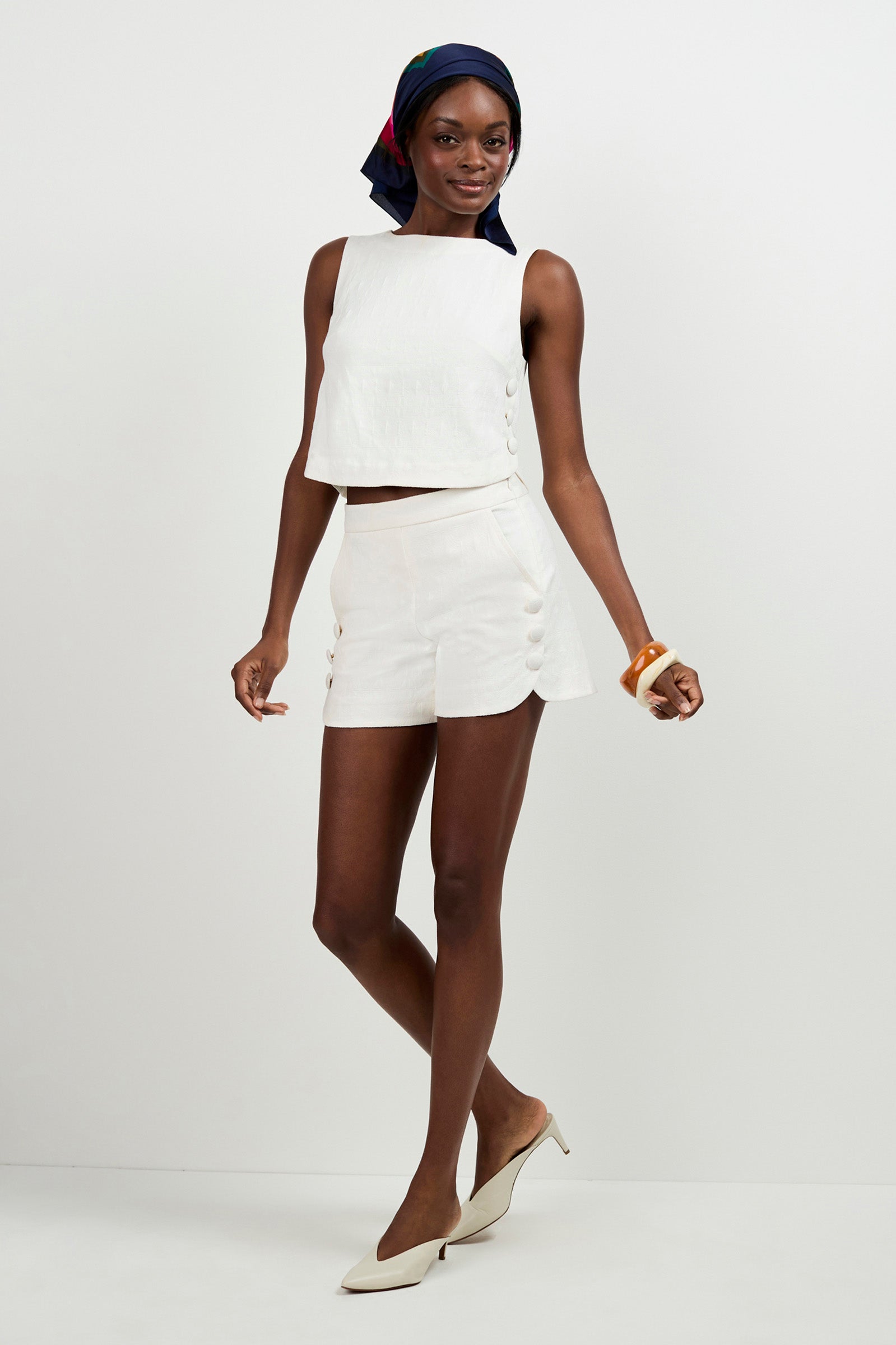 A woman poses against a white background wearing the Duomo Button Side Shorts, a sleeveless white crop top, beige heels, a dark headscarf, and a chunky bracelet.