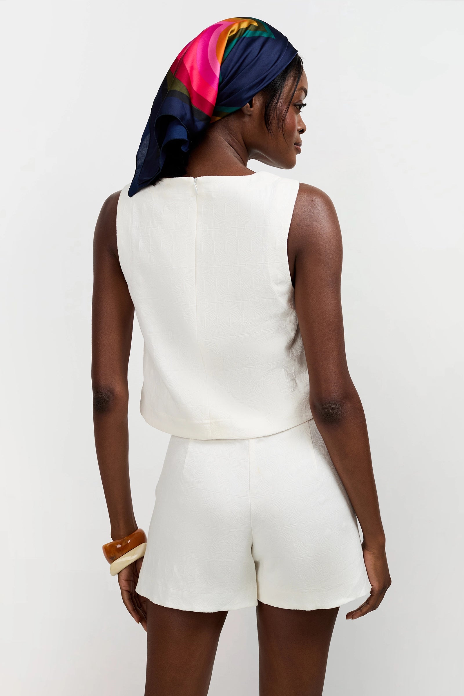 A woman stands with her back to the camera in a Siara Button Side Top and matching shorts, accessorized with a colorful silk scarf and chunky bangles. The background is plain white.
