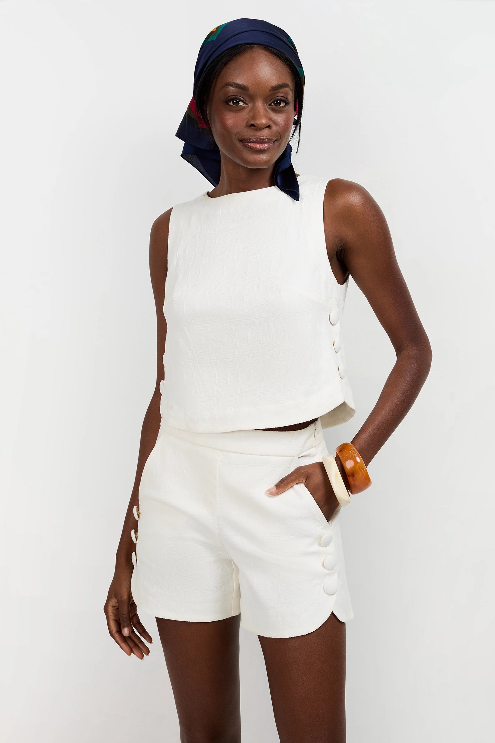 A woman stands against a plain white background, smiling slightly, wearing the Siara Button Side Top— a sleeveless textured white top—paired with matching high-waisted shorts, a navy headscarf, and a chunky brown bracelet.