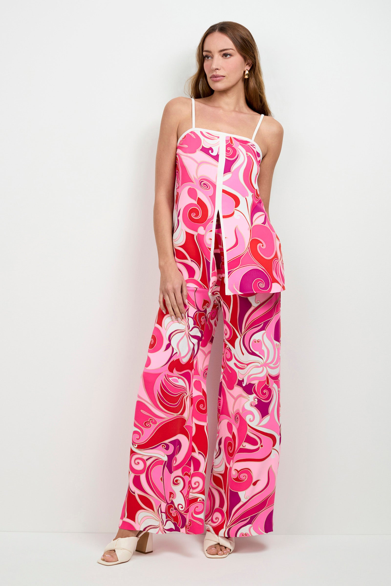 A woman poses against a white wall wearing the Aliza Tunic Top—a pink and red swirled-patterned sleeveless top—paired with matching wide-leg pants and white heeled sandals, her hair down, one hand on her hip.