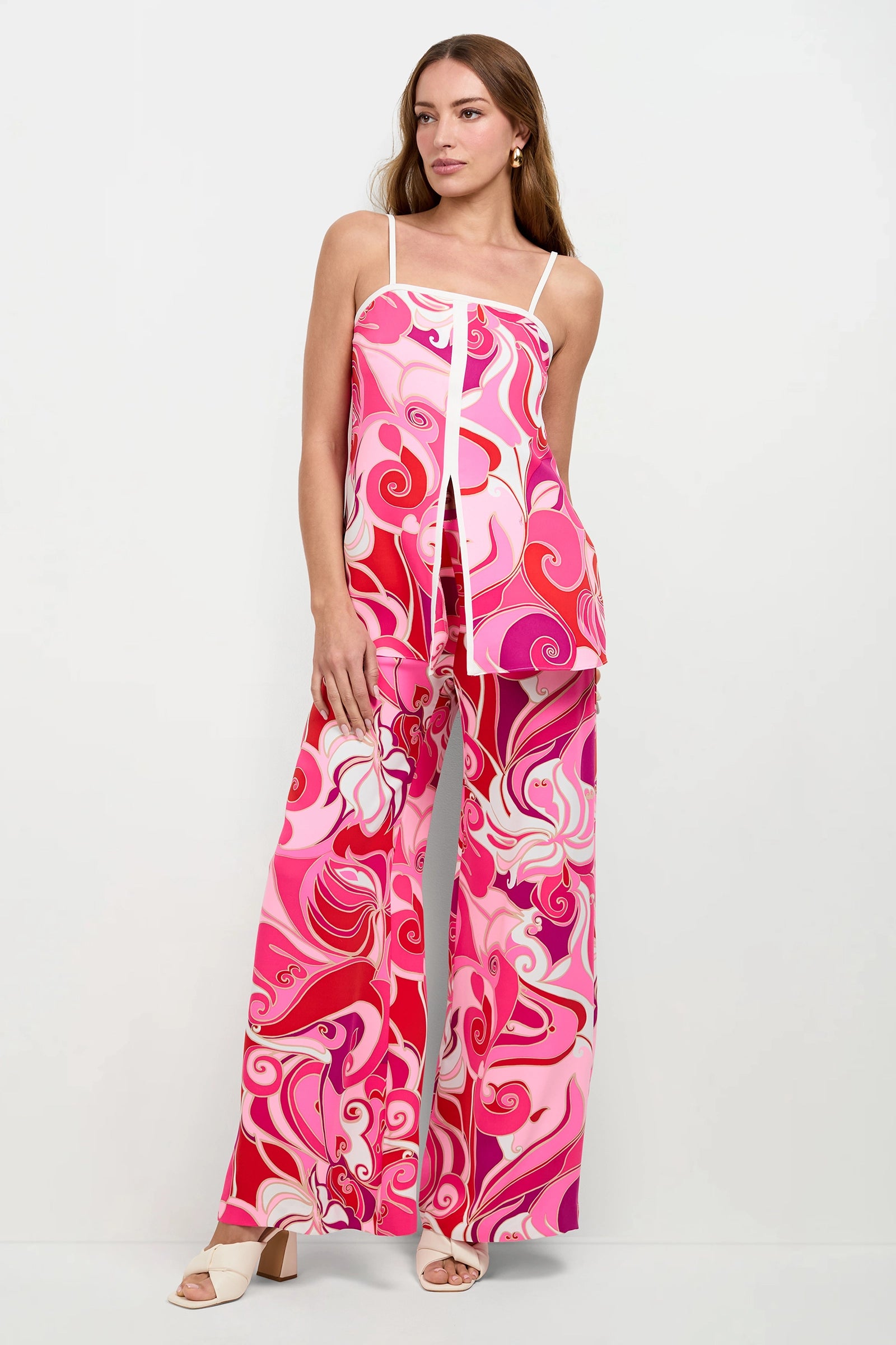 A woman stands against a plain background, wearing the Aliza Tunic Top—featuring a vibrant pink and red swirl pattern with white trim—paired with matching wide-leg pants, cream heeled sandals, and minimal jewelry.