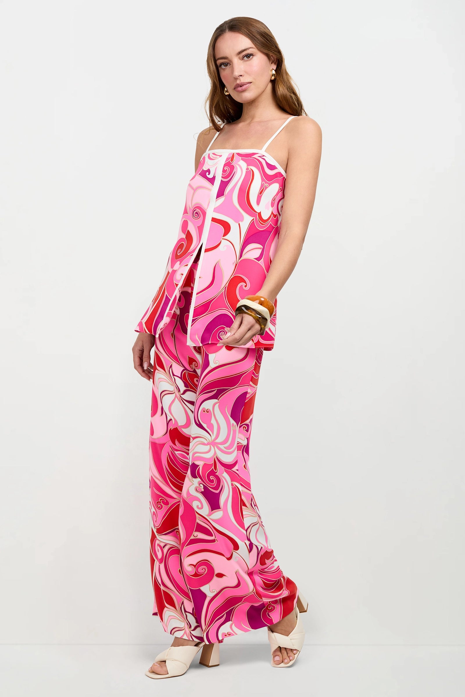 A woman poses in the Aliza Tunic Top, a sleeveless piece with pink, red, and white swirling patterns. She pairs it with matching wide-leg pants, white heels, gold earrings, and chunky bracelets against a plain white background.
