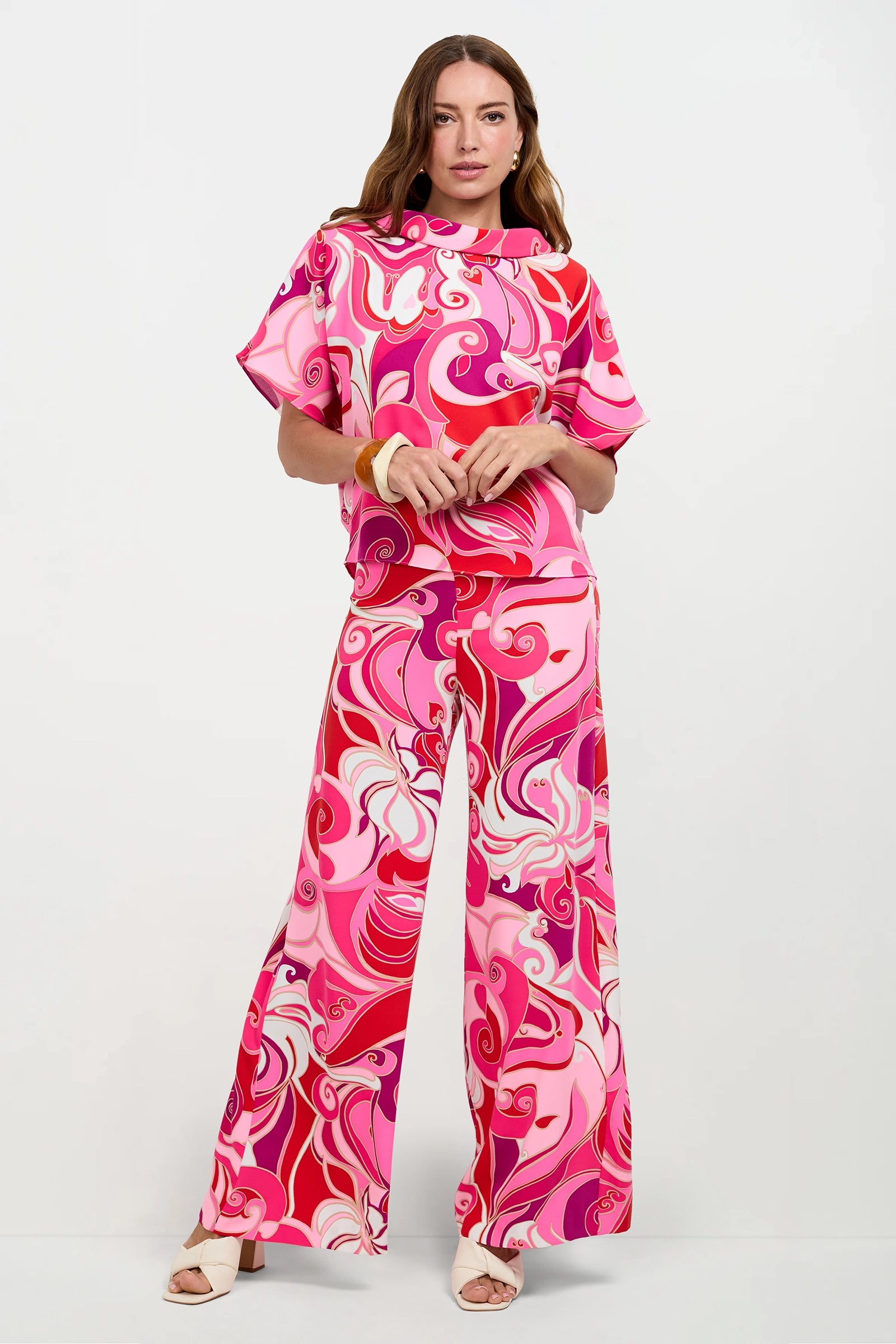 A woman wears the Honeysuckle Boatneck Top in Darsena, styled with matching wide-leg pants. The set features a colorful, retro swirl pattern in pink, red, and white. She completes the look with white heels and a yellow bracelet.