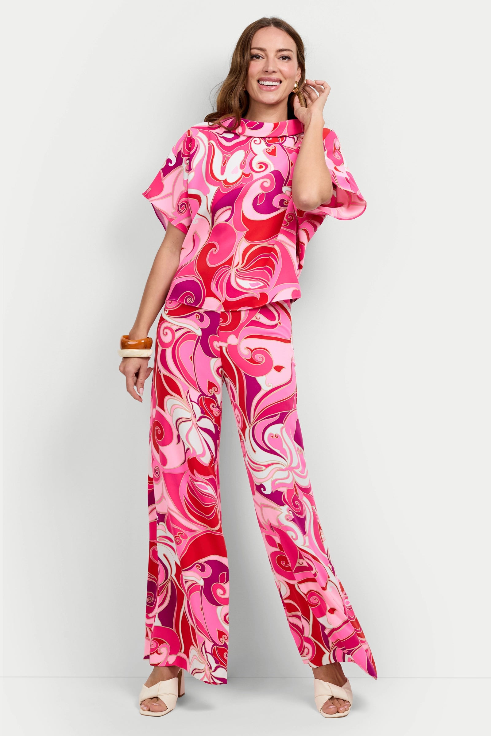 A woman with long hair smiles while wearing the Honeysuckle Boatneck Top and matching wide-leg pants featuring a vibrant pink, red, and white swirl pattern. She completes her look with open-toe shoes and a chunky orange bracelet.