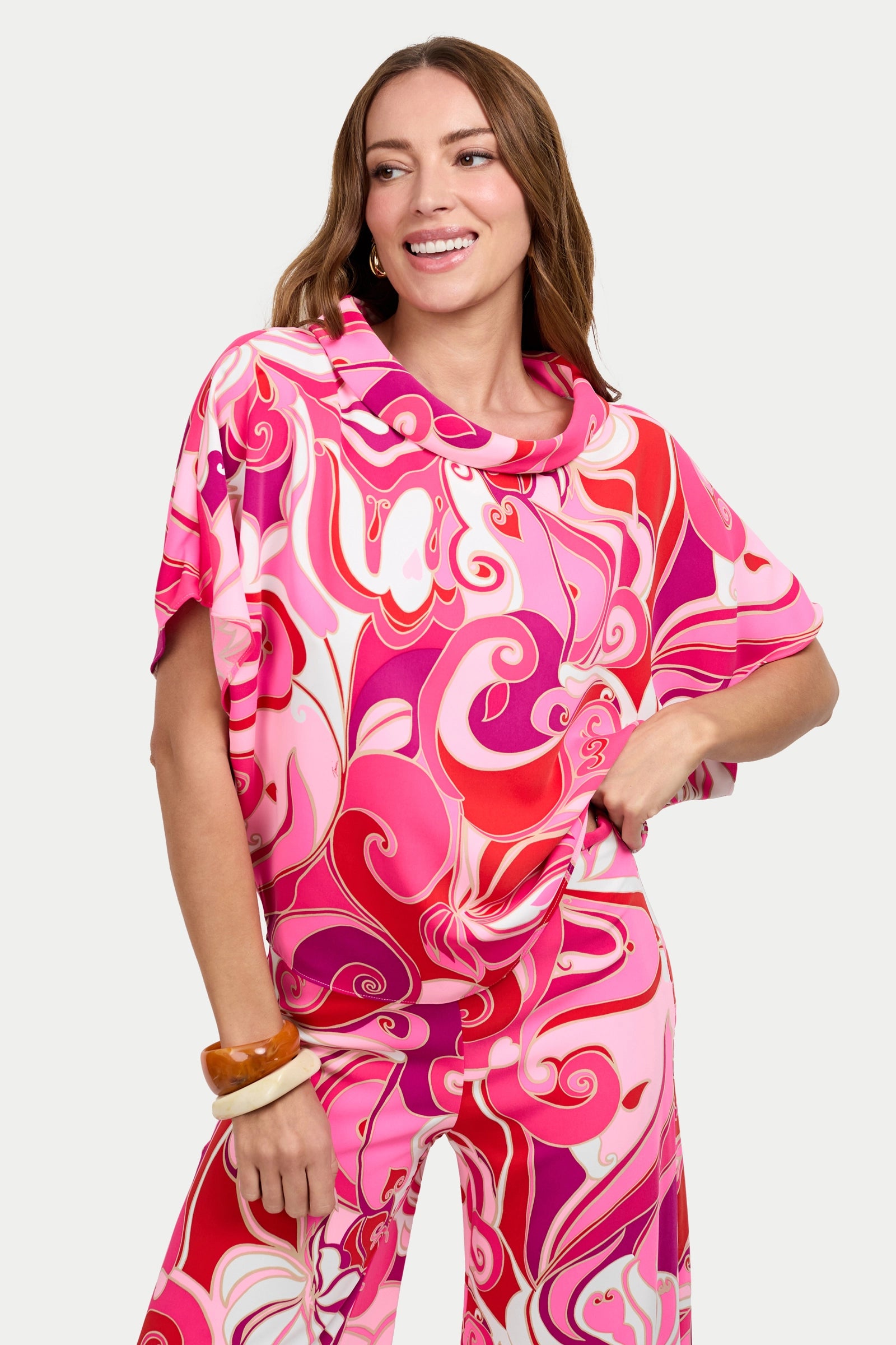 A woman with light brown hair smiles while wearing the Honeysuckle Boatneck Top, featuring a vibrant pink and red swirl pattern. She pairs it with matching pants and chunky bracelets on her wrist for a stylish look.