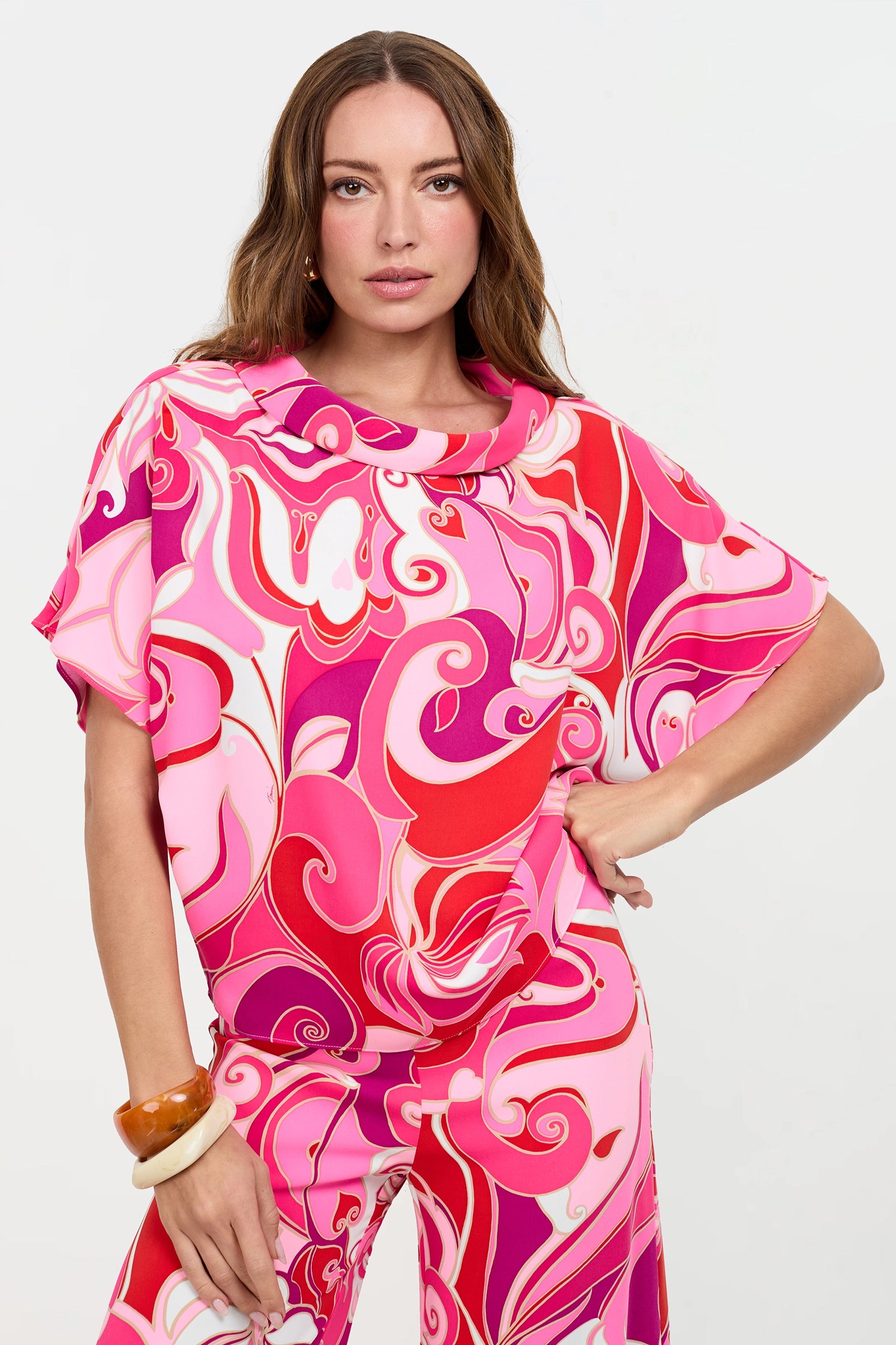 A woman with long brown hair poses against a plain white background, wearing the Honeysuckle Boatneck Top featuring a vibrant pink and red swirling pattern. She completes the look with matching pants and large bracelets on her wrist.
