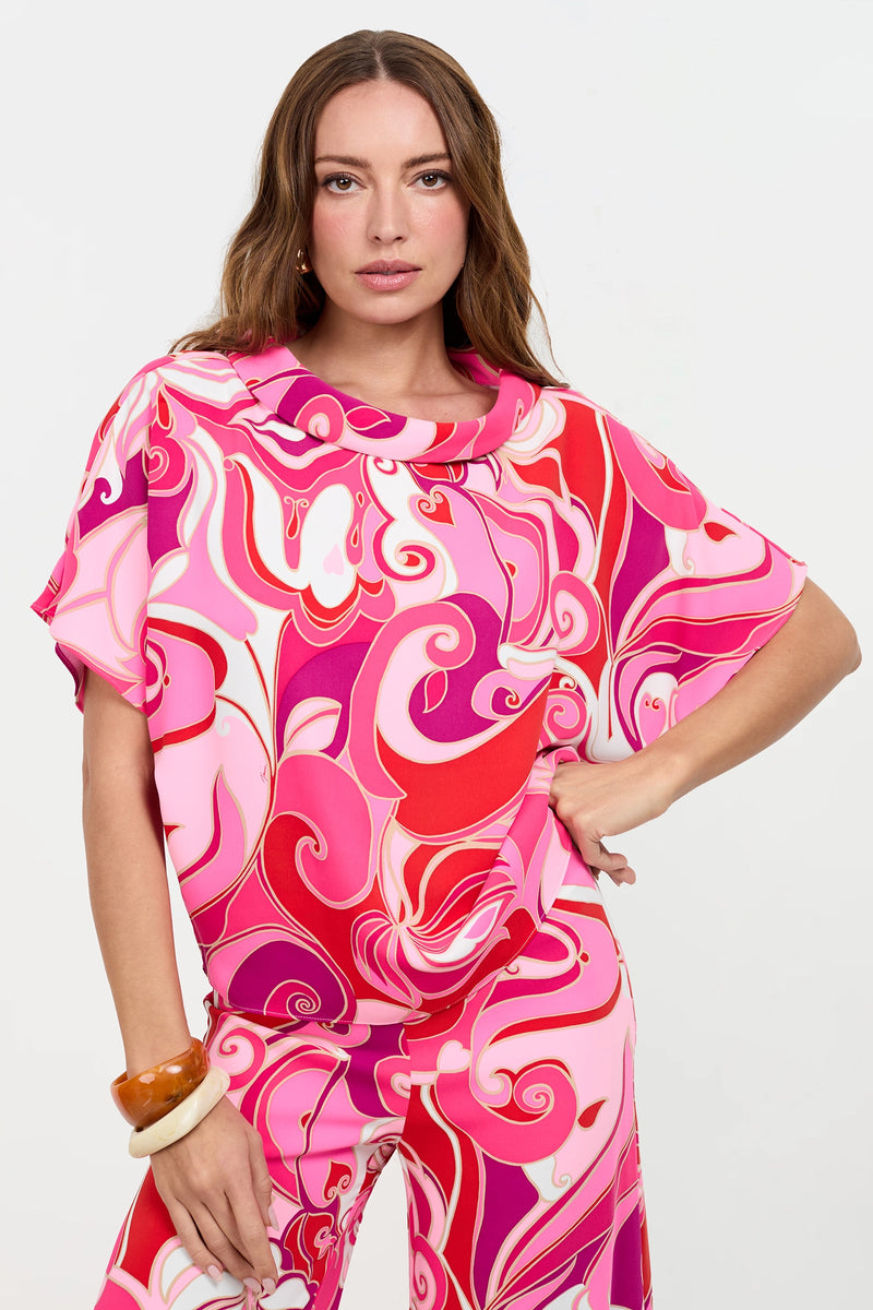 A woman with long brown hair poses against a plain white background, wearing the Honeysuckle Boatneck Top featuring a vibrant pink and red swirling pattern. She completes the look with matching pants and large bracelets on her wrist.