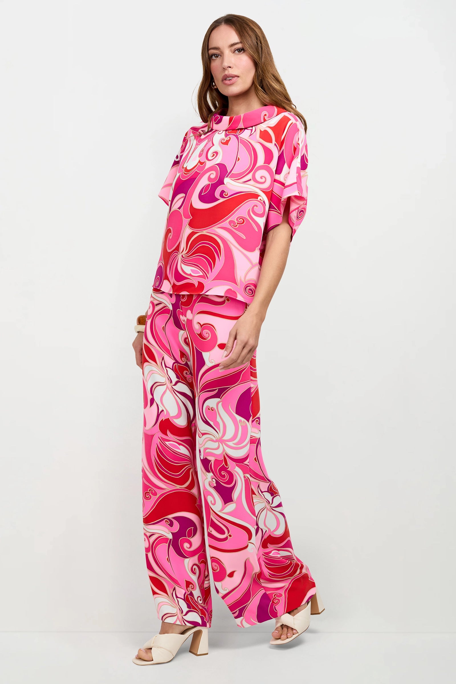A woman stands against a white background wearing the Honeysuckle Boatneck Top with matching pink, red, and white psychedelic swirl wide-leg pants and white open-toe heels.