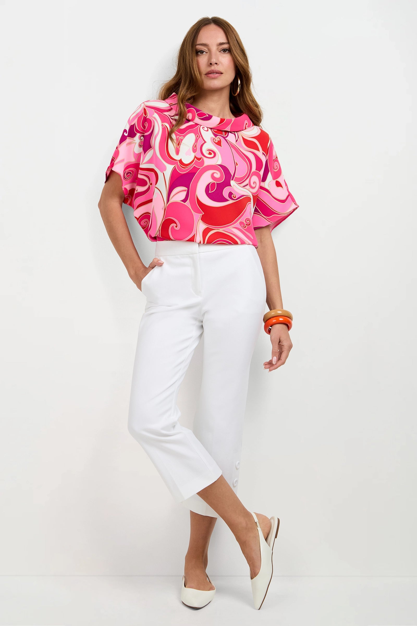 A woman poses against a white background wearing the Honeysuckle Boatneck Top in Darsena, paired with white cropped pants, white pointed-toe flats, and orange bracelets.