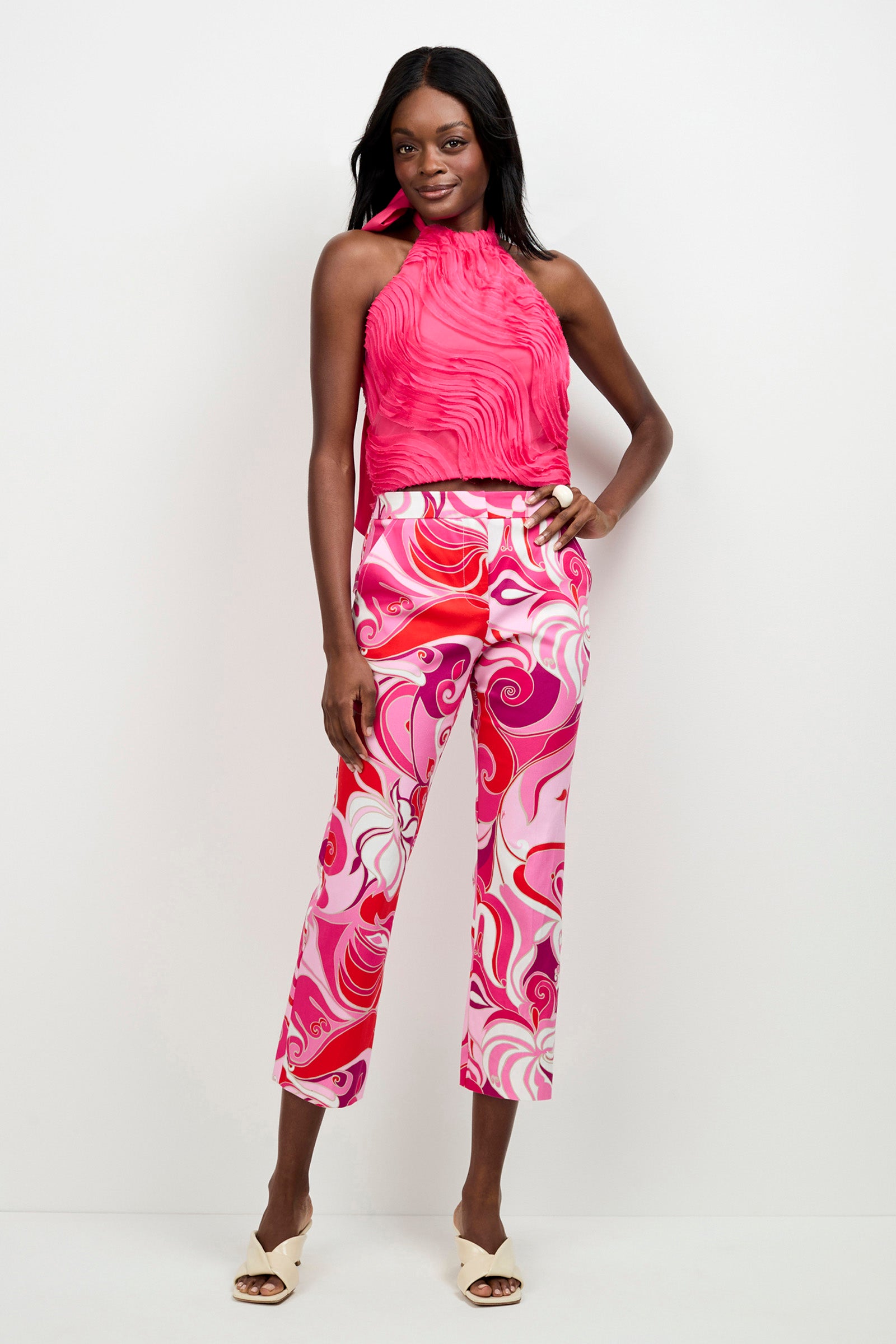 A woman stands against a white background wearing the Myka Organza Halter Top, paired with high-waisted, pink-white-red swirl patterned pants and white open-toe heels. She smiles slightly.