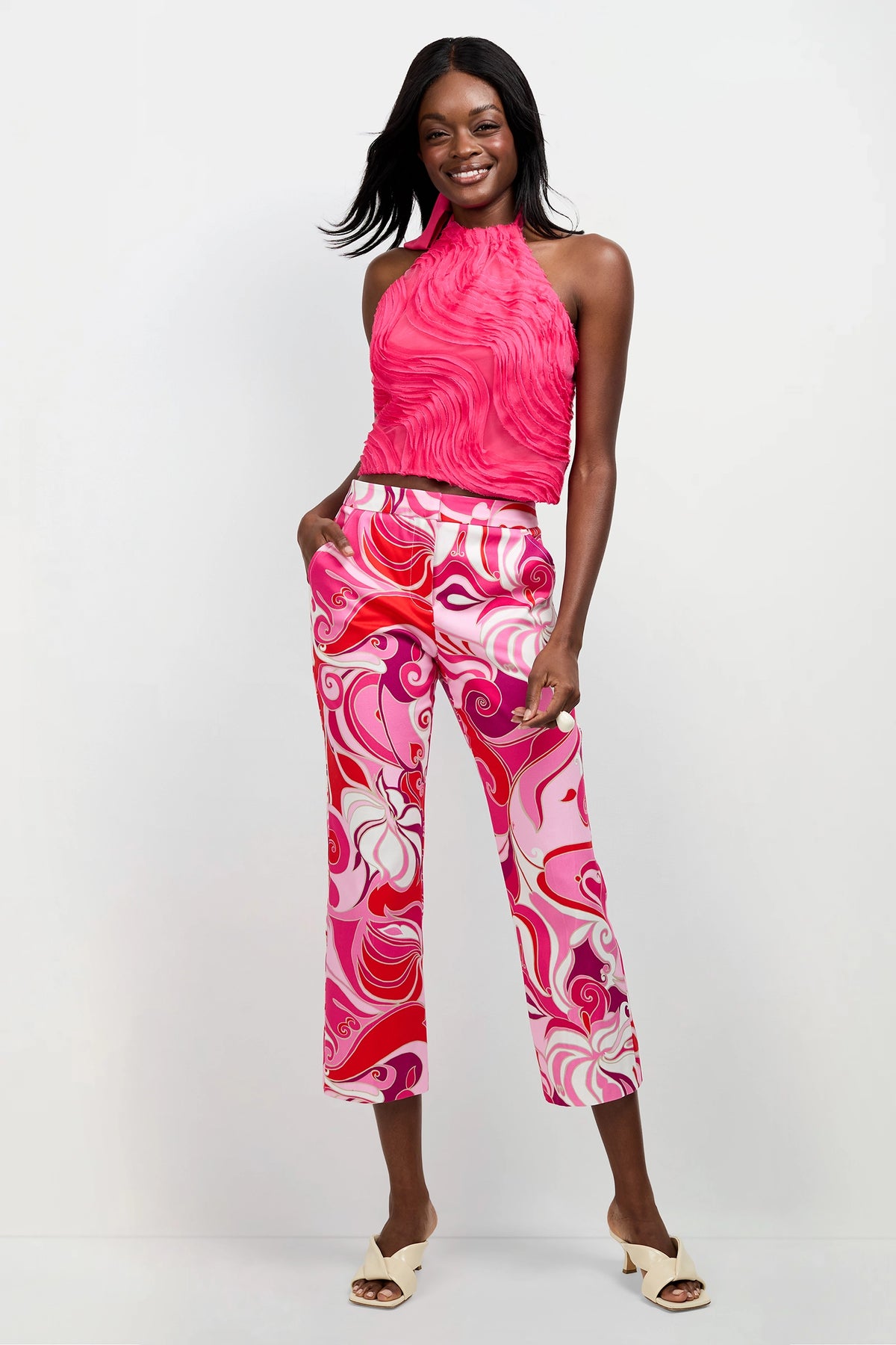 A woman smiles against a white background, wearing the Myka Organza Halter Top in Magenta Milano, colorful pink and red swirl-patterned pants, and white open-toe heeled sandals.