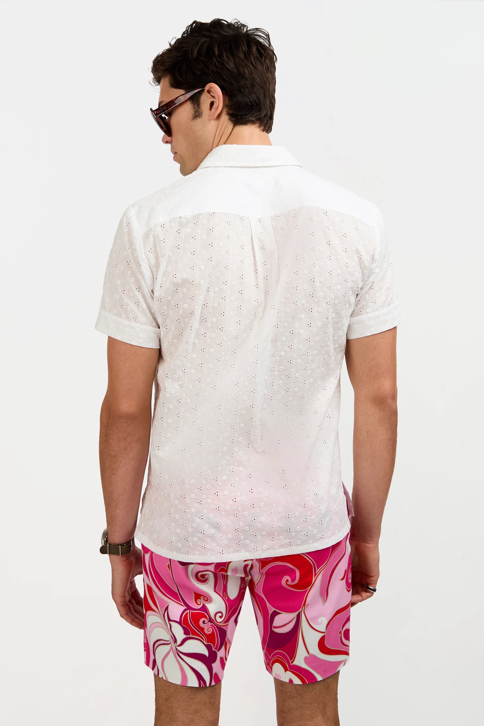 A man with dark hair, sunglasses, and a watch stands against a light background wearing the Branson Short-Sleeve Shirt and bright pink, white, and red patterned shorts.
