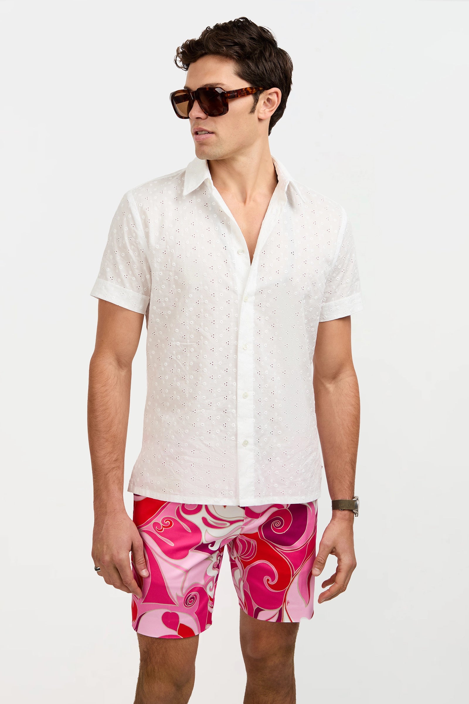 A man stands against a plain white background wearing the Branson Short-Sleeve Shirt, large sunglasses, and pink patterned shorts.