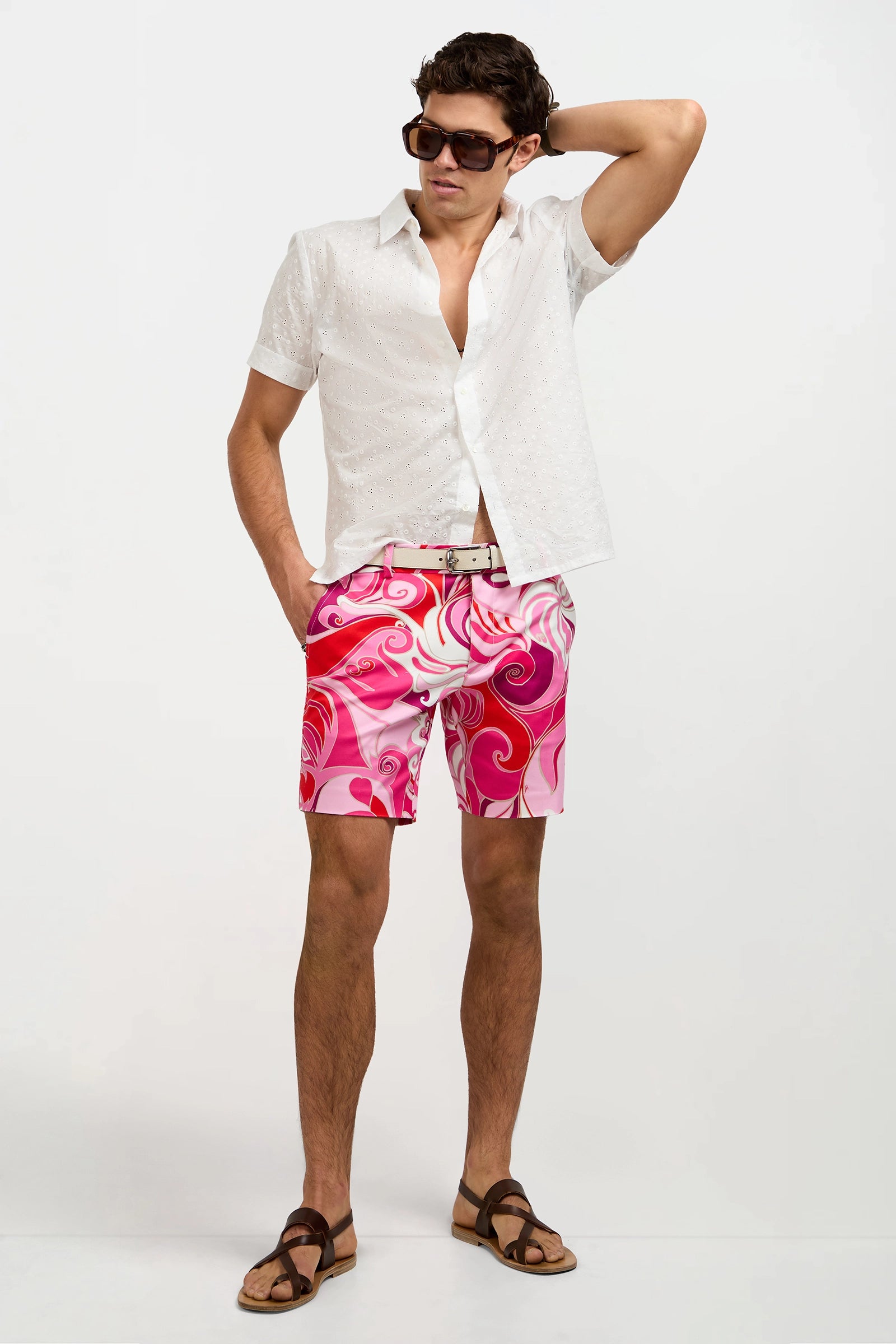 A man in dark sunglasses, brown sandals, a white belt, pink and white swirl shorts, and a Branson Short-Sleeve Shirt in White stands with one hand behind his head against a plain white background.