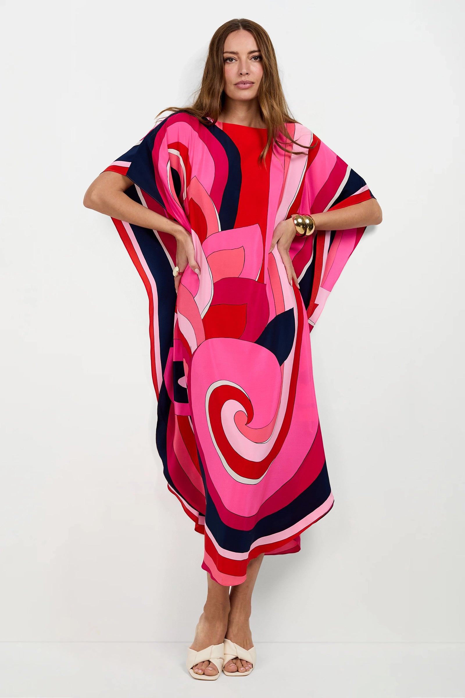 A woman with long brown hair poses against a white background in the Jalani Maxi Caftan Dress, featuring bold swirling pink, red, and navy patterns. She wears neutral sandals and a gold bracelet, with her hands on her hips.