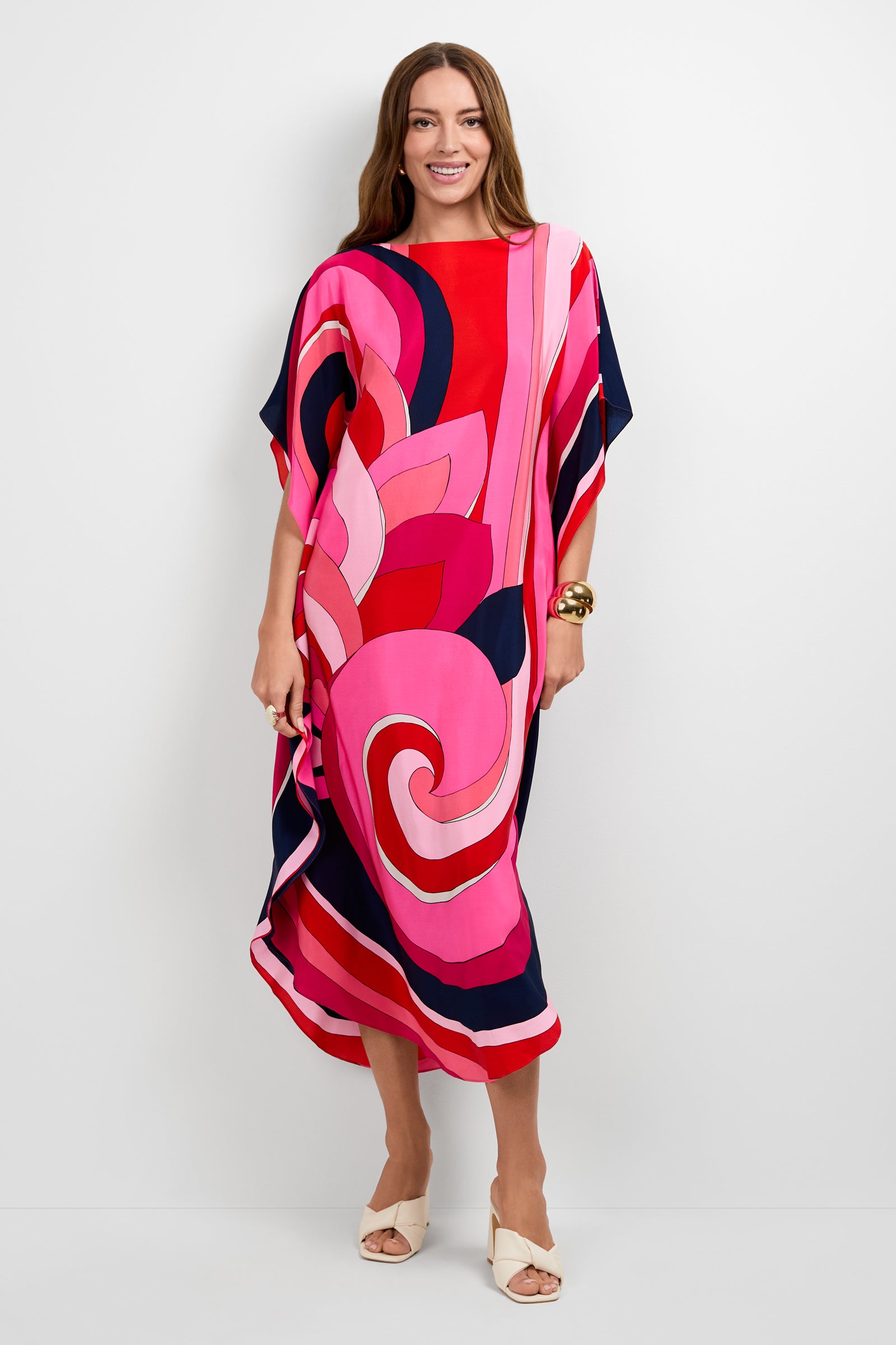 A woman smiles in the Jalani Maxi Caftan Dress, featuring bright, flowing fabric with bold red, pink, and navy swirls. She pairs it with white open-toe heels and a gold bracelet while posing against a plain white background.