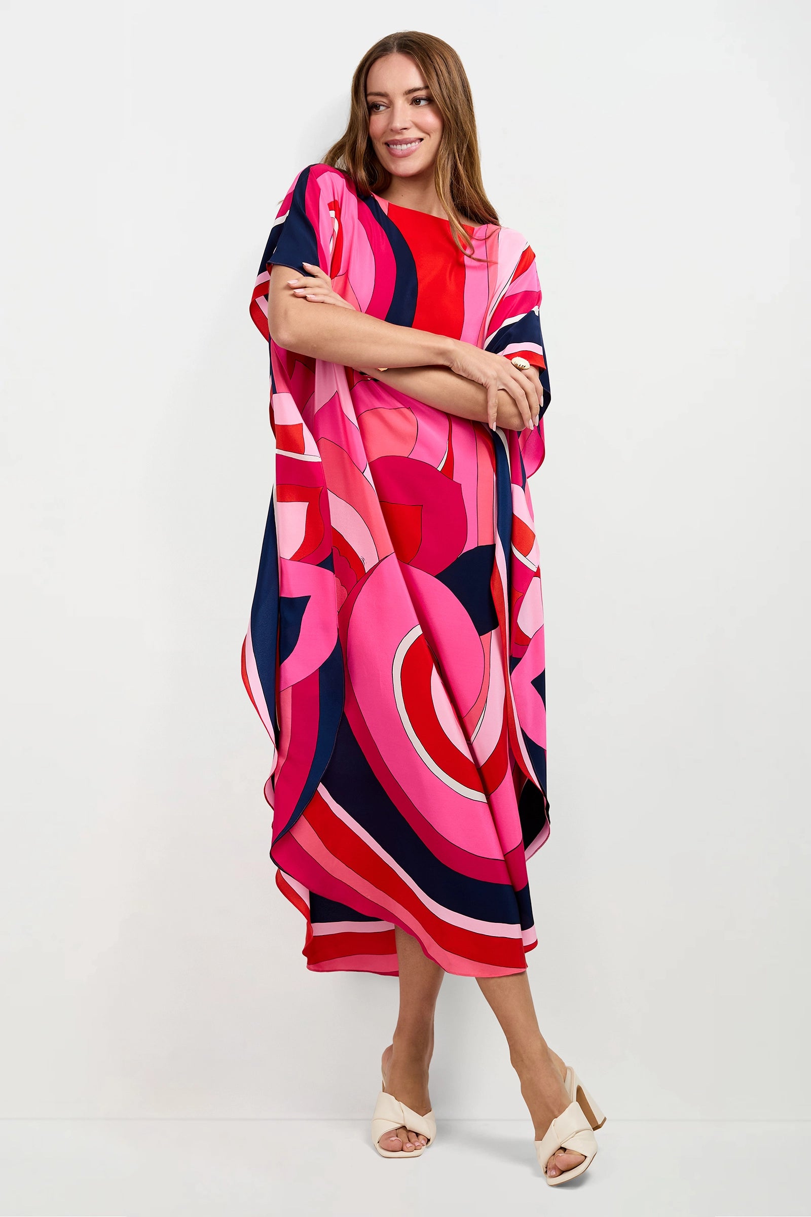 A woman stands against a white wall, smiling, wearing the Jalani Maxi Caftan Dress featuring bold pink, red, navy, and white geometric patterns with cream open-toe heels.