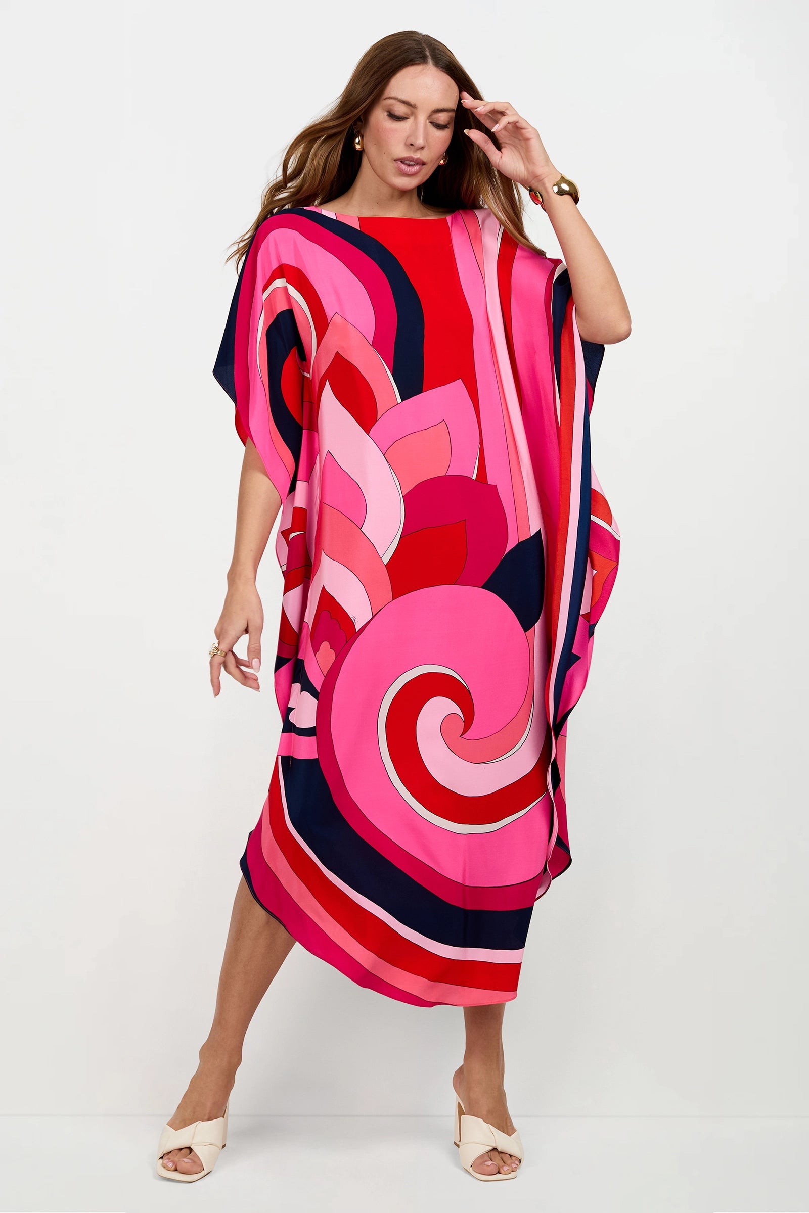A woman models the Jalani Maxi Caftan Dress, a loose, brightly colored dress with bold pink, red, and navy swirling patterns. She stands against a plain white background with one hand near her head and the other by her side.