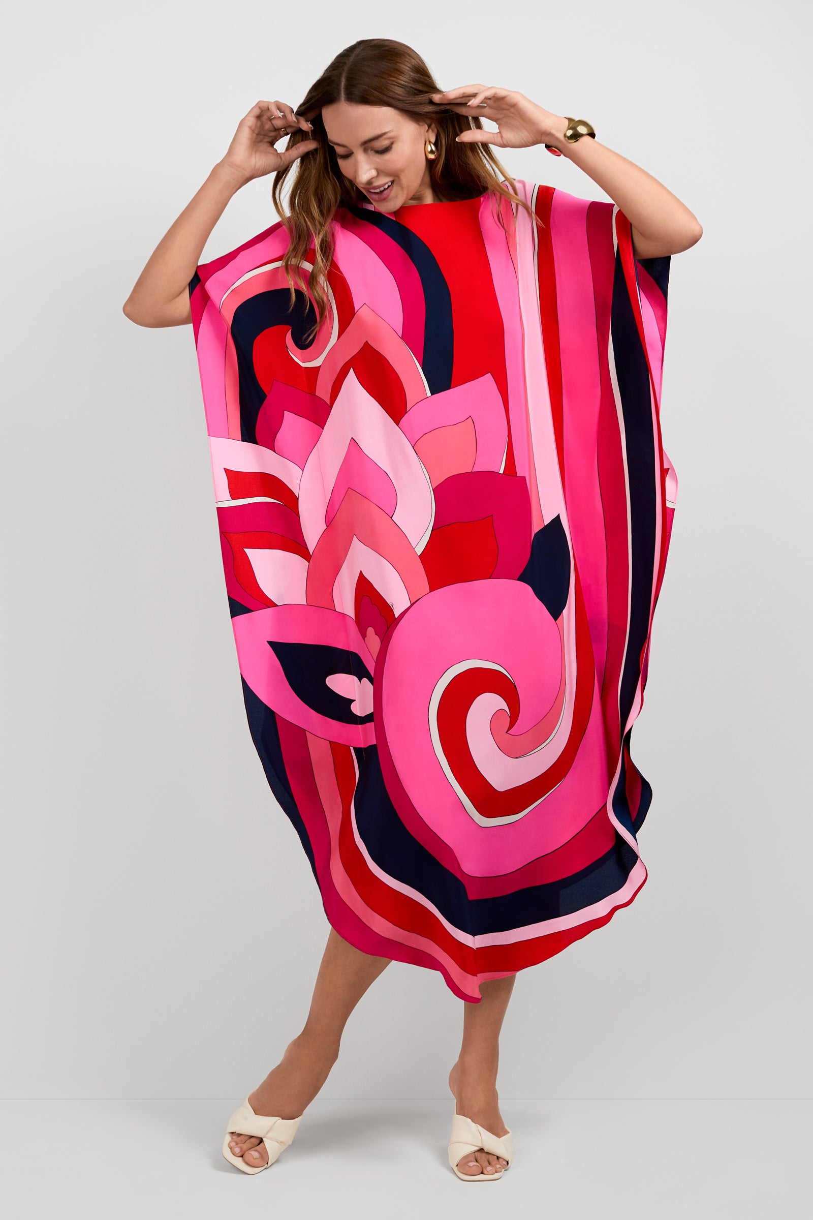 A smiling woman wears the Jalani Maxi Caftan Dress, featuring bold pink, red, and navy abstract patterns. She stands against a plain background with arms raised playfully, paired with white open-toe heels and gold earrings.