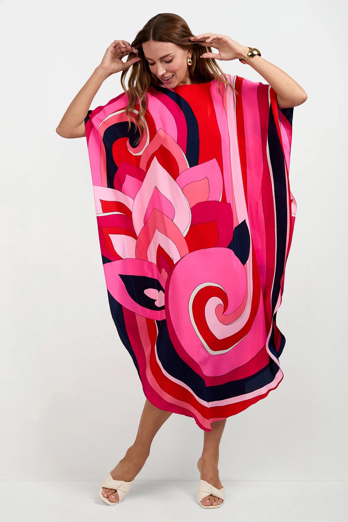 A woman smiles barefoot against a white background, wearing the Jalani Maxi Caftan Dress—a loose, colorful piece with bold pink, red, navy, and white abstract patterns.