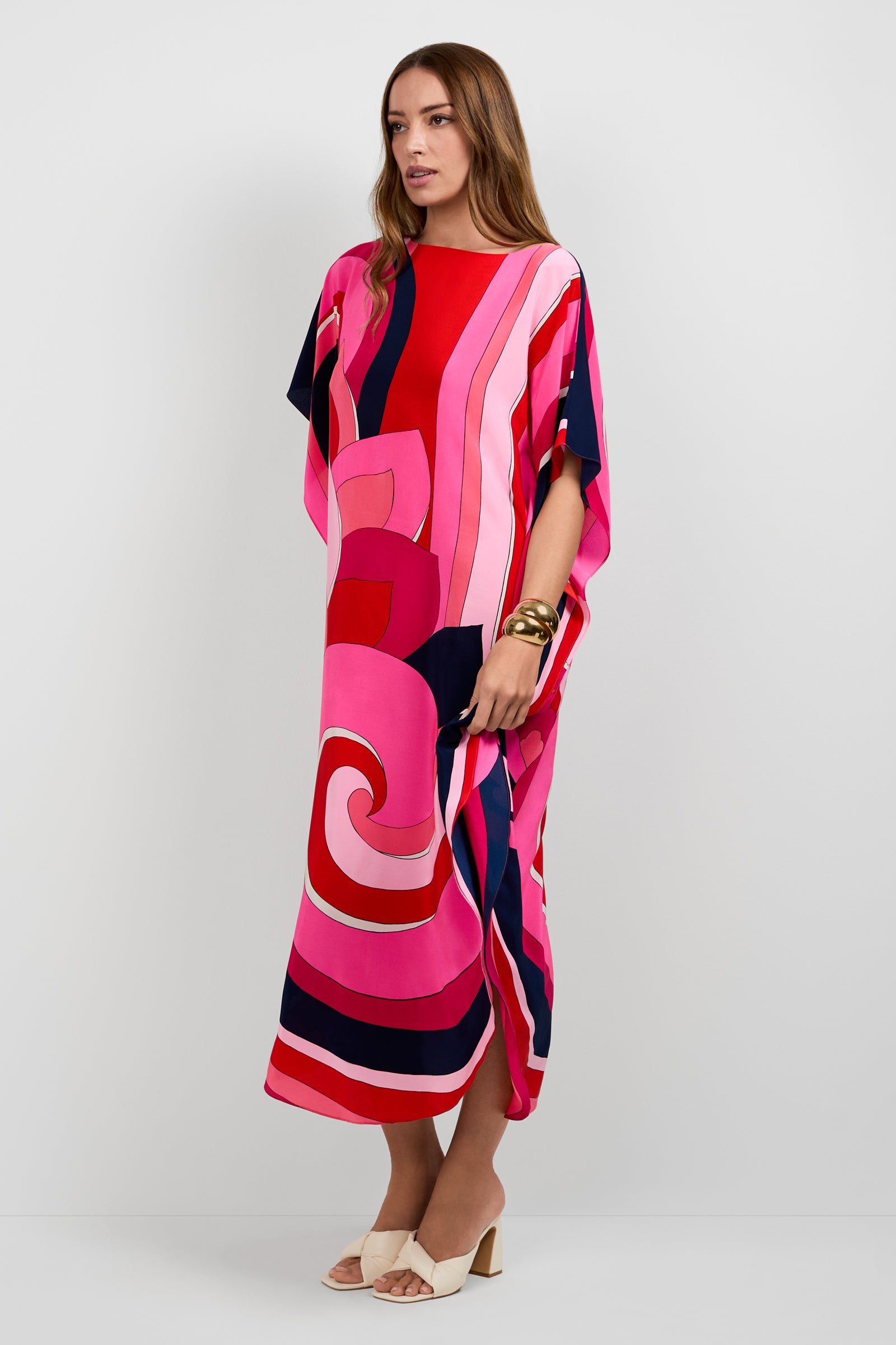 A woman models the Jalani Maxi Caftan Dress, featuring bold abstract red, pink, navy, and white patterns. She has long brown hair and pairs the dress with white heeled sandals and gold bangles against a plain white background.