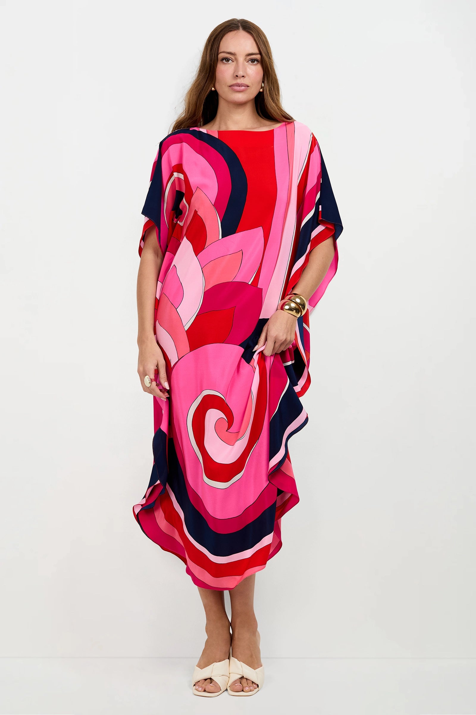 A woman with long brown hair wears the Jalani Maxi Caftan Dress, a loose, knee-length piece featuring bold pink, red, navy, and white abstract patterns. She pairs it with white open-toe shoes and a chunky gold bracelet against a white background.
