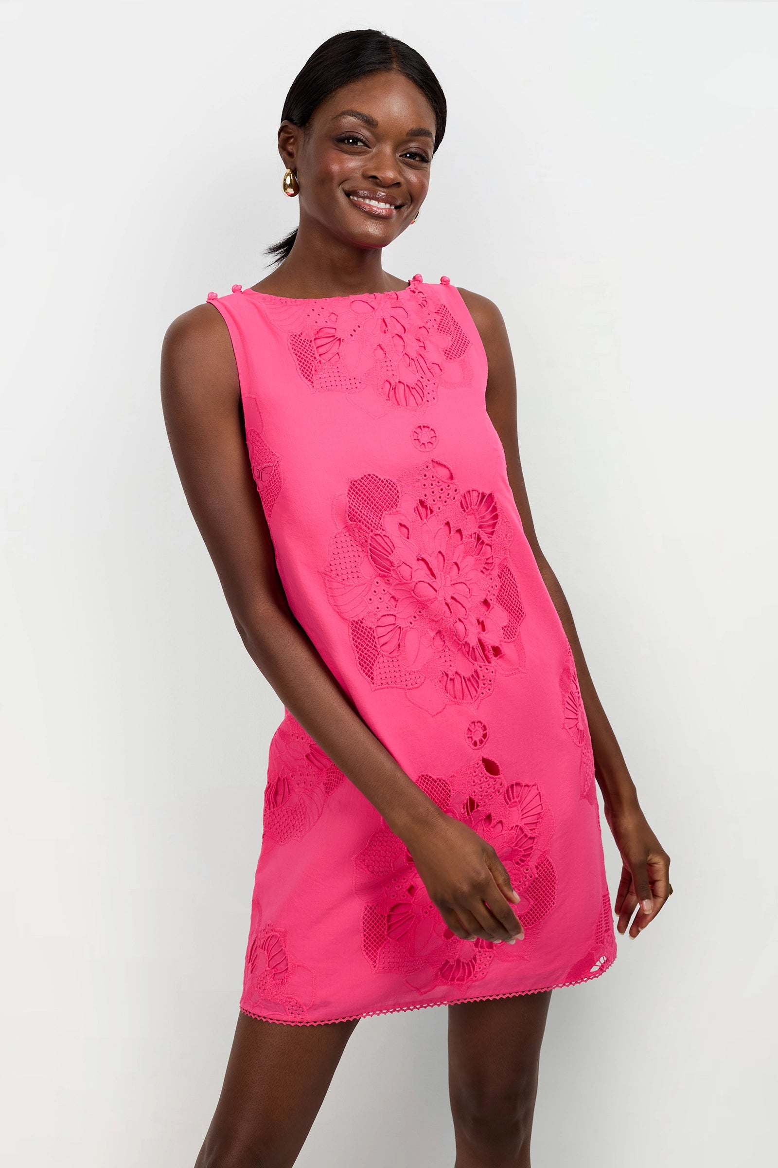 A woman smiles while wearing the Francesca Shift Dress, a sleeveless bright pink piece with floral cutout patterns. She stands against a plain white background, one hand by her side and the other gently touching her hip.
