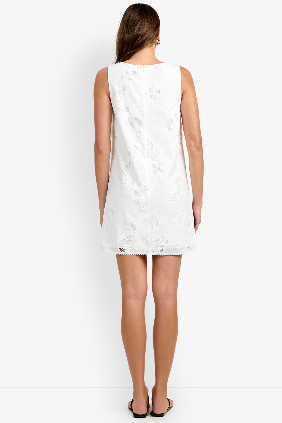 A woman stands facing away, wearing the sleeveless Francesca Shift Dress—a short white dress with a subtle floral pattern—paired with flat sandals, against a plain white background.
