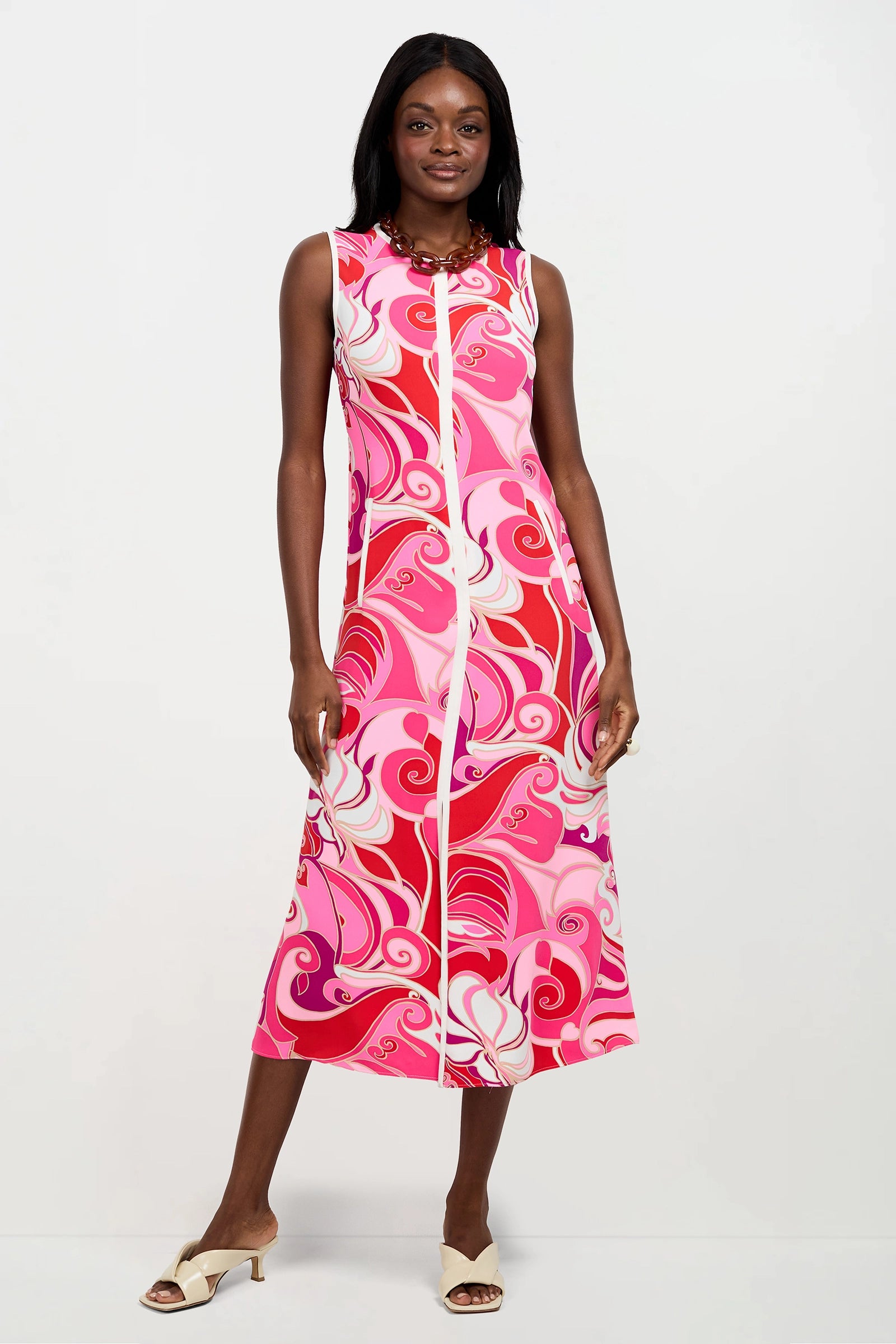 A woman poses against a white background wearing the Magnifique Sleeveless Dress, an ankle-length piece featuring a vibrant pink, red, and white swirl print. She completes the look with white heeled sandals.