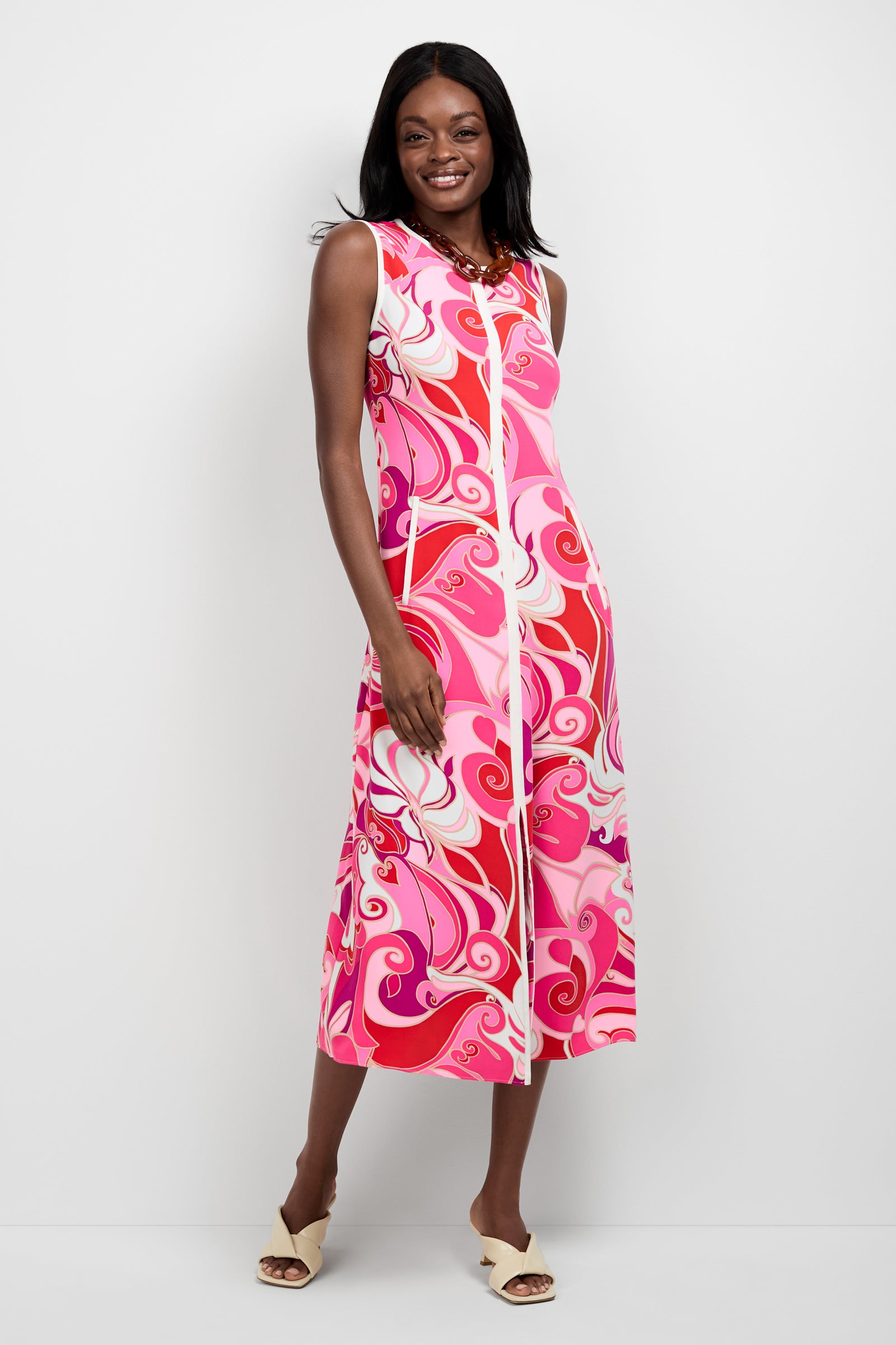 A woman smiles in the Magnifique Sleeveless Dress, featuring a bold pink, red, and white swirl pattern. The calf-length dress is paired with cream open-toed heels. She stands before a plain white background.