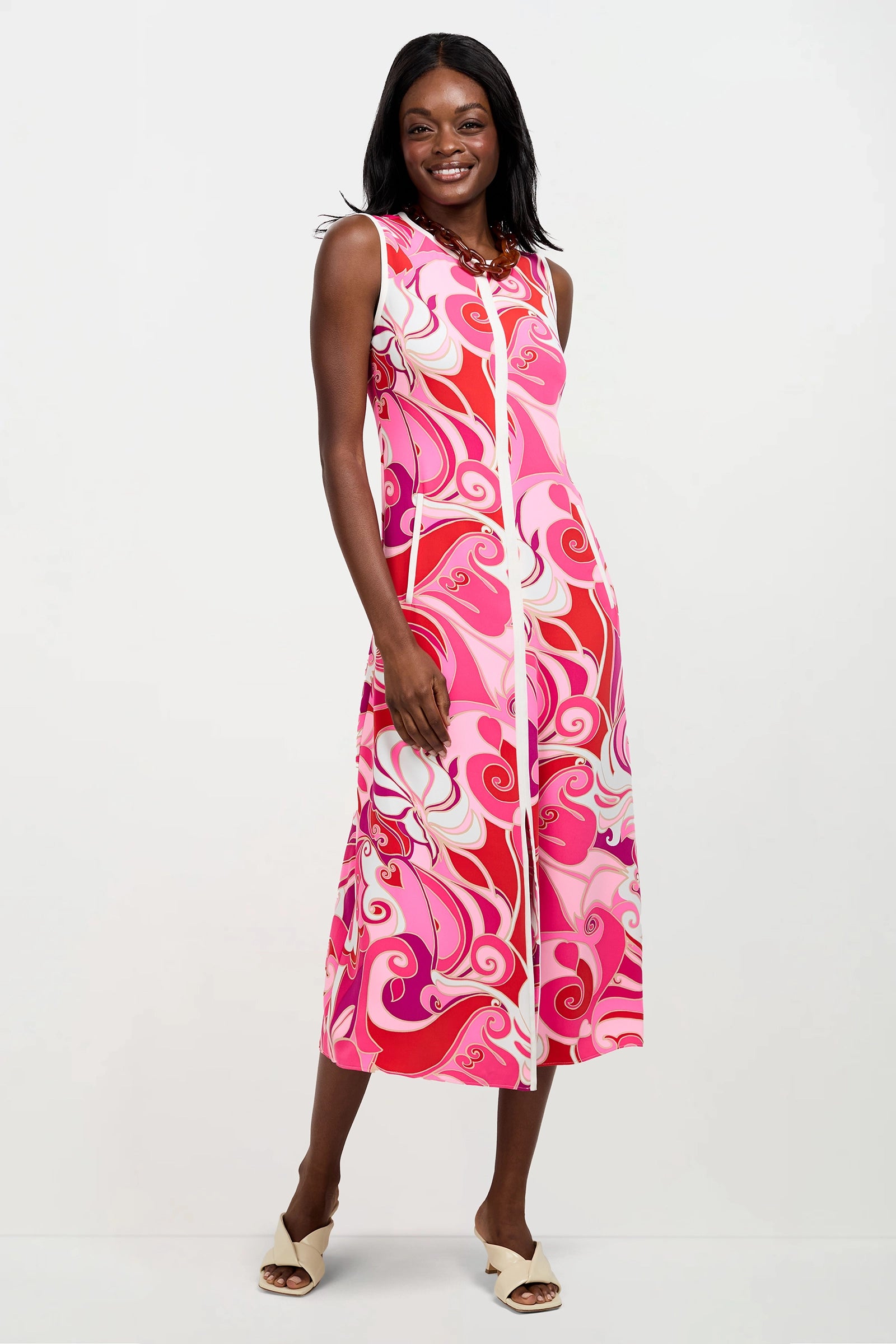 A woman smiles in the Magnifique Sleeveless Dress, an ankle-length piece with a bold pink, white, and red swirly pattern. She pairs it with light open-toe shoes and poses against a plain white background.