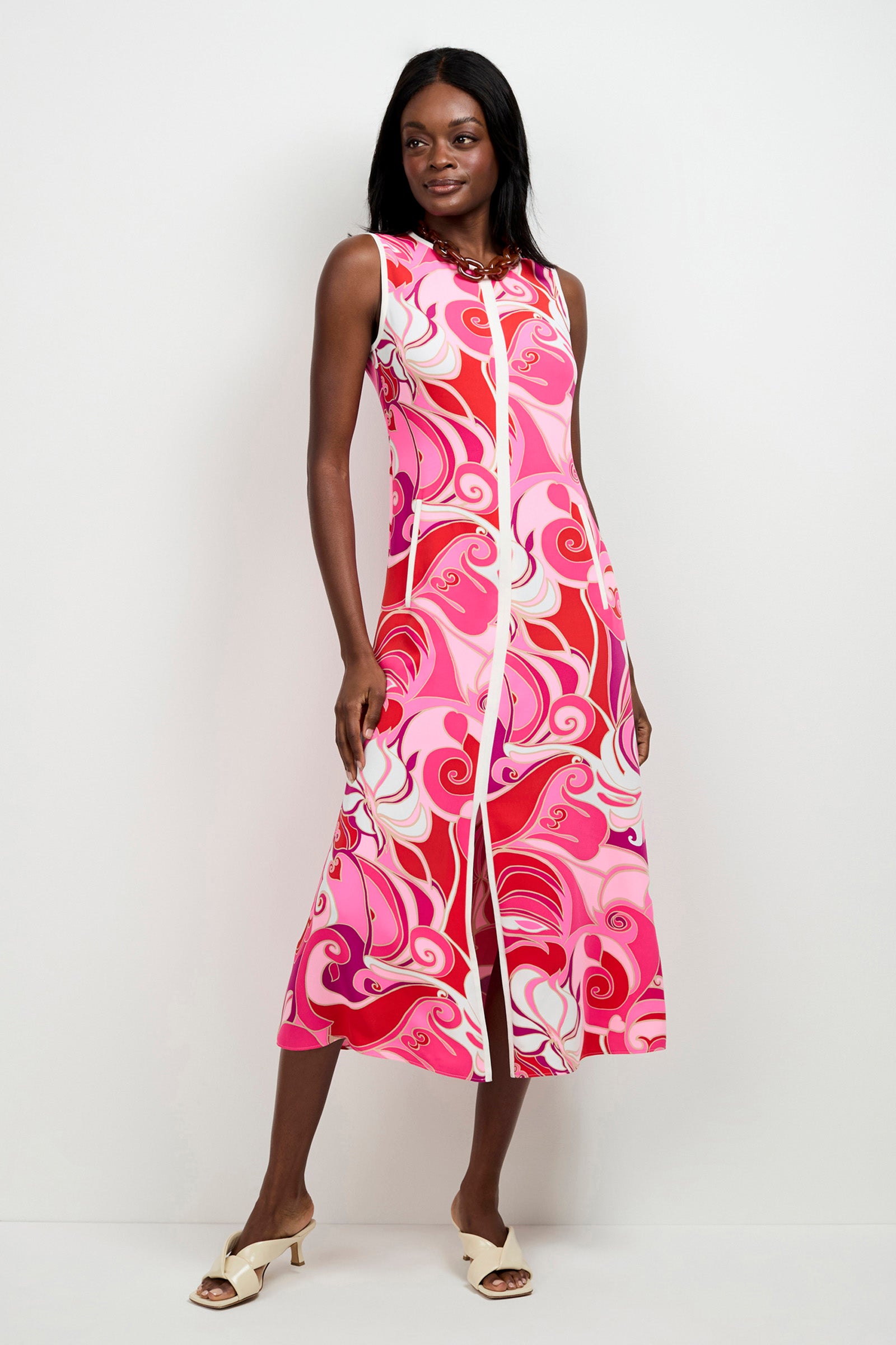 A woman stands against a white wall wearing the Magnifique Sleeveless Dress, an ankle-length style with bold pink, red, and white swirls. She completes the look with cream heeled sandals and a necklace.