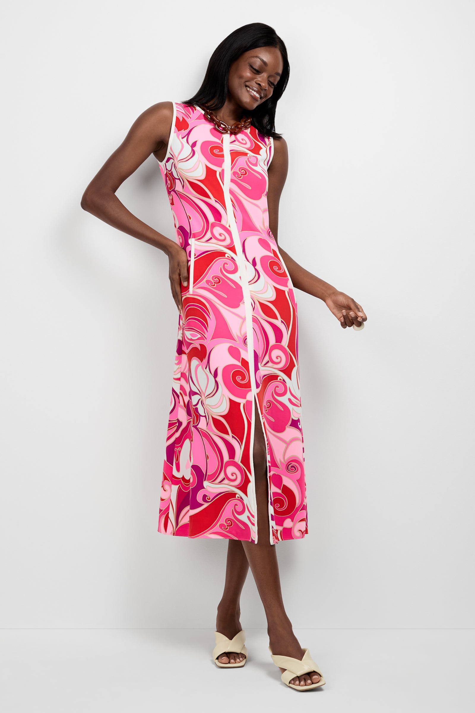 A woman stands against a white background, smiling and wearing the Magnifique Sleeveless Dress—a pink and white patterned midi with a front slit. She has one hand on her hip, the other holding the skirt, paired with cream-colored sandals.