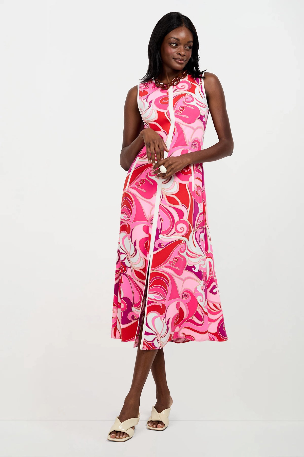 A woman wears the Magnifique Sleeveless Dress, featuring a knee-length cut and a vibrant pink, red, and white swirling pattern. She stands confidently with a smile, one leg forward, and pairs the dress with light open-toe shoes.