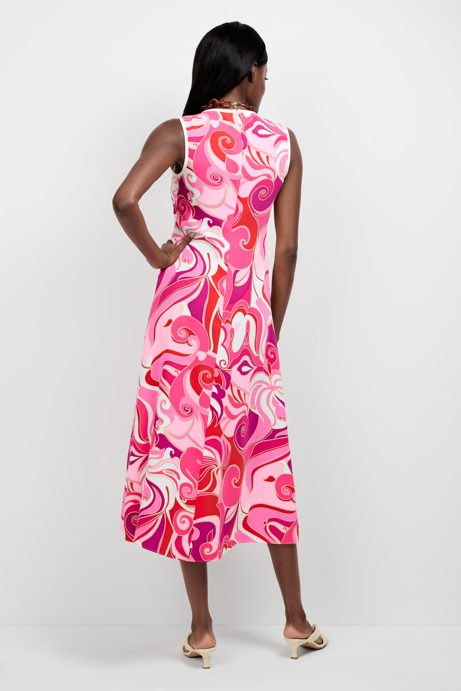 A woman with dark hair, seen from behind, wears the Magnifique Sleeveless Dress—an ankle-length dress featuring a vibrant pink, red, and white swirling pattern—paired with light-colored heeled sandals.